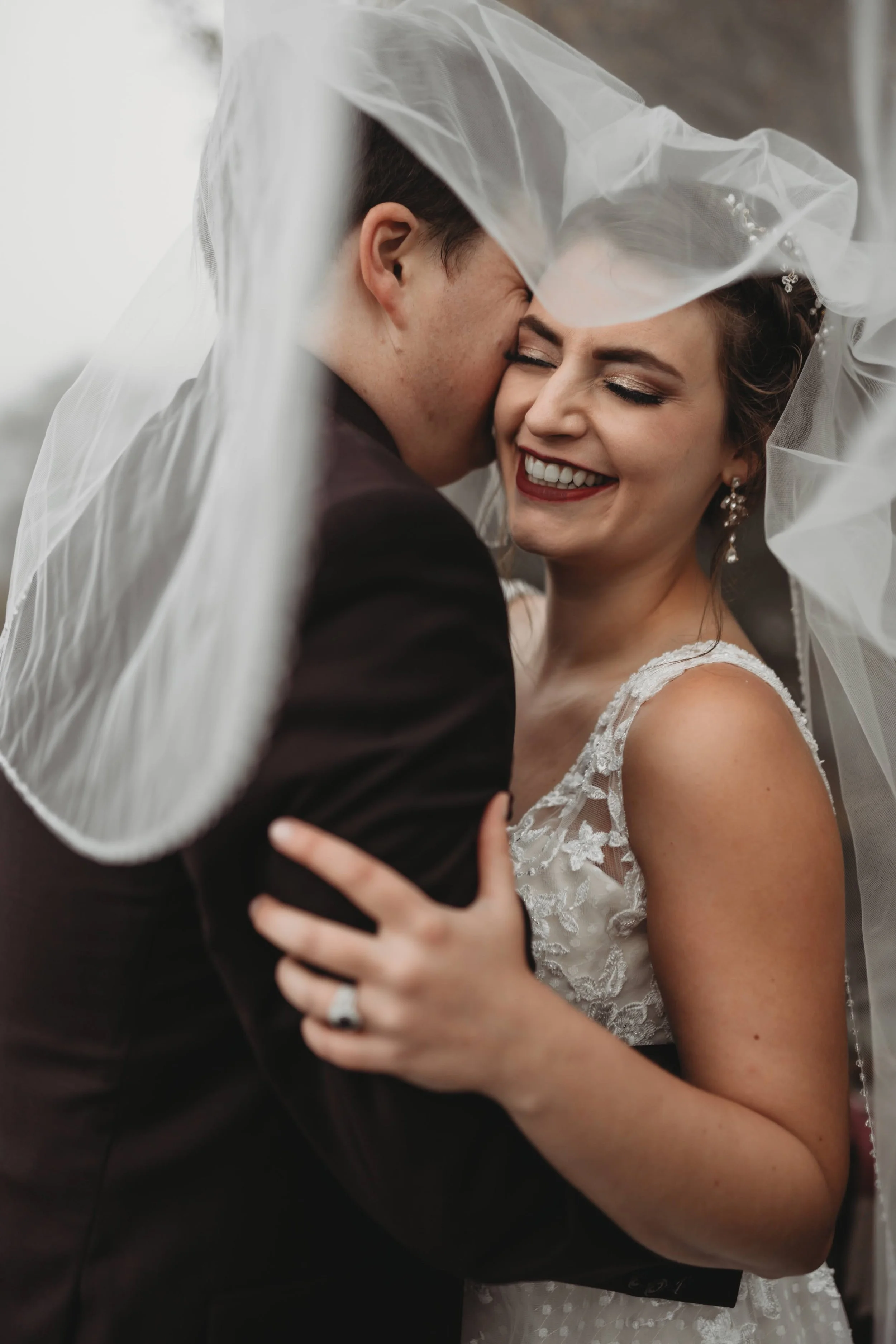 Bride smiles while embracing groom under her veil in an intimate wedding portrait.