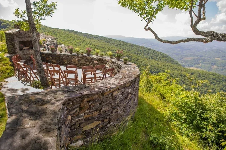 Outdoor ceremony setup with wooden chairs on a curved stone terrace overlooking forested mountains.