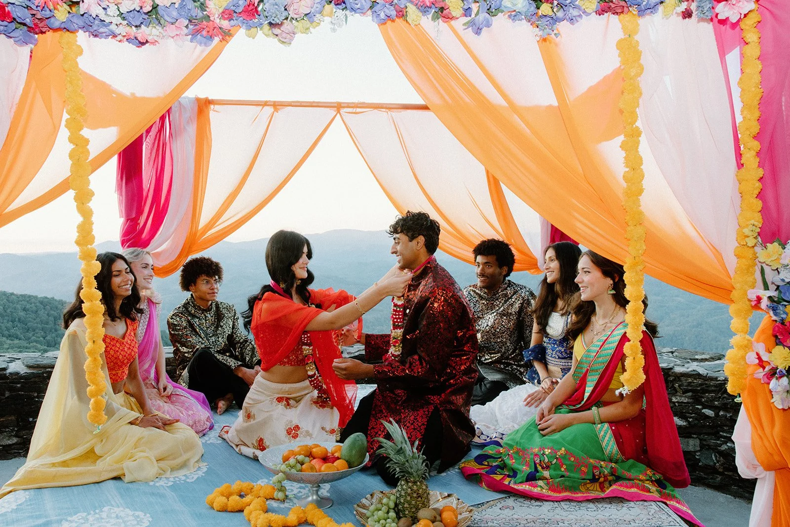 Couple sits under a colorful draped mandap with friends around them, with mountains visible beyond the ceremony space.