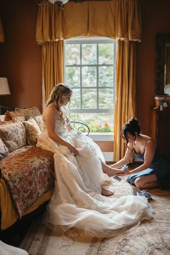 Bride getting ready as a bridesmaid helps with her shoes beside a window.