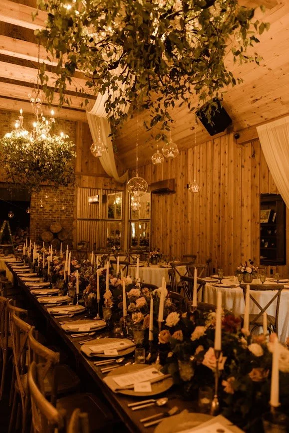 Warm, candlelit barn wedding reception with long banquet tables, wooden walls, and soft draped fabric overhead.