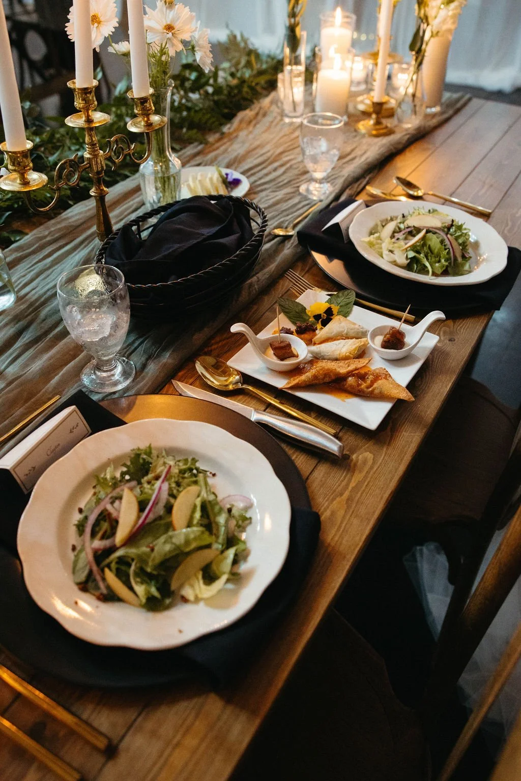 Candlelit rustic dinner table with plated green salads, black napkins, and a shared appetizer platter