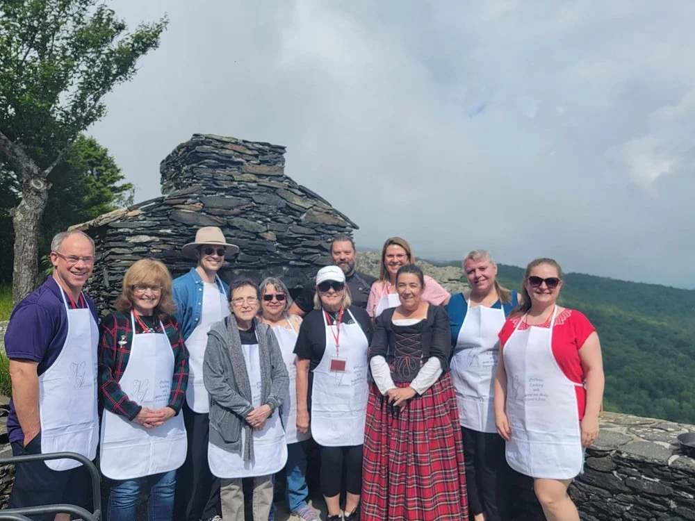 A group of people wearing white aprons stand in front of a stone structure with a mountain view in the background.