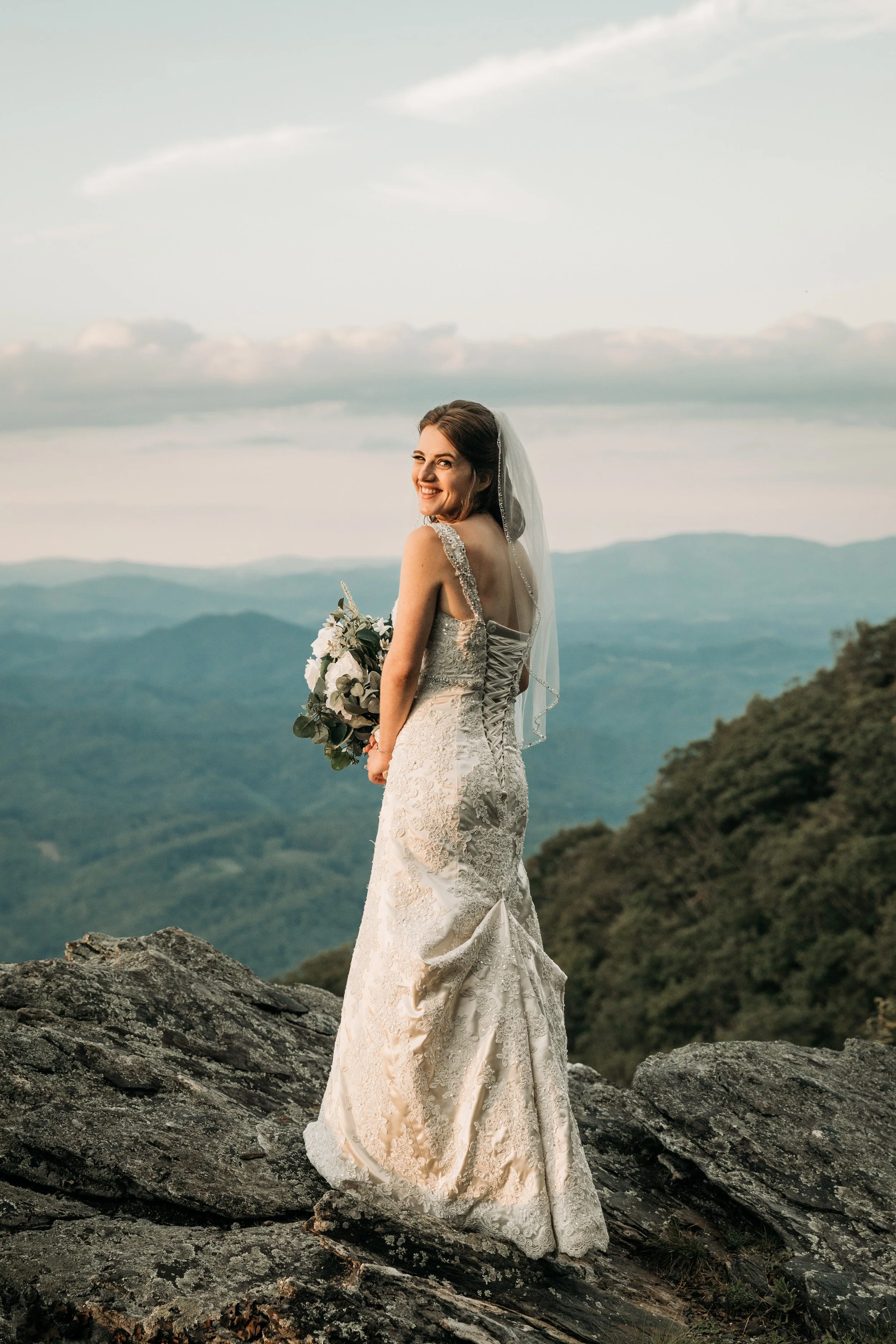 Bride looking over her shoulder on a rocky mountain overlook while holding a white and greenery bouquet.
