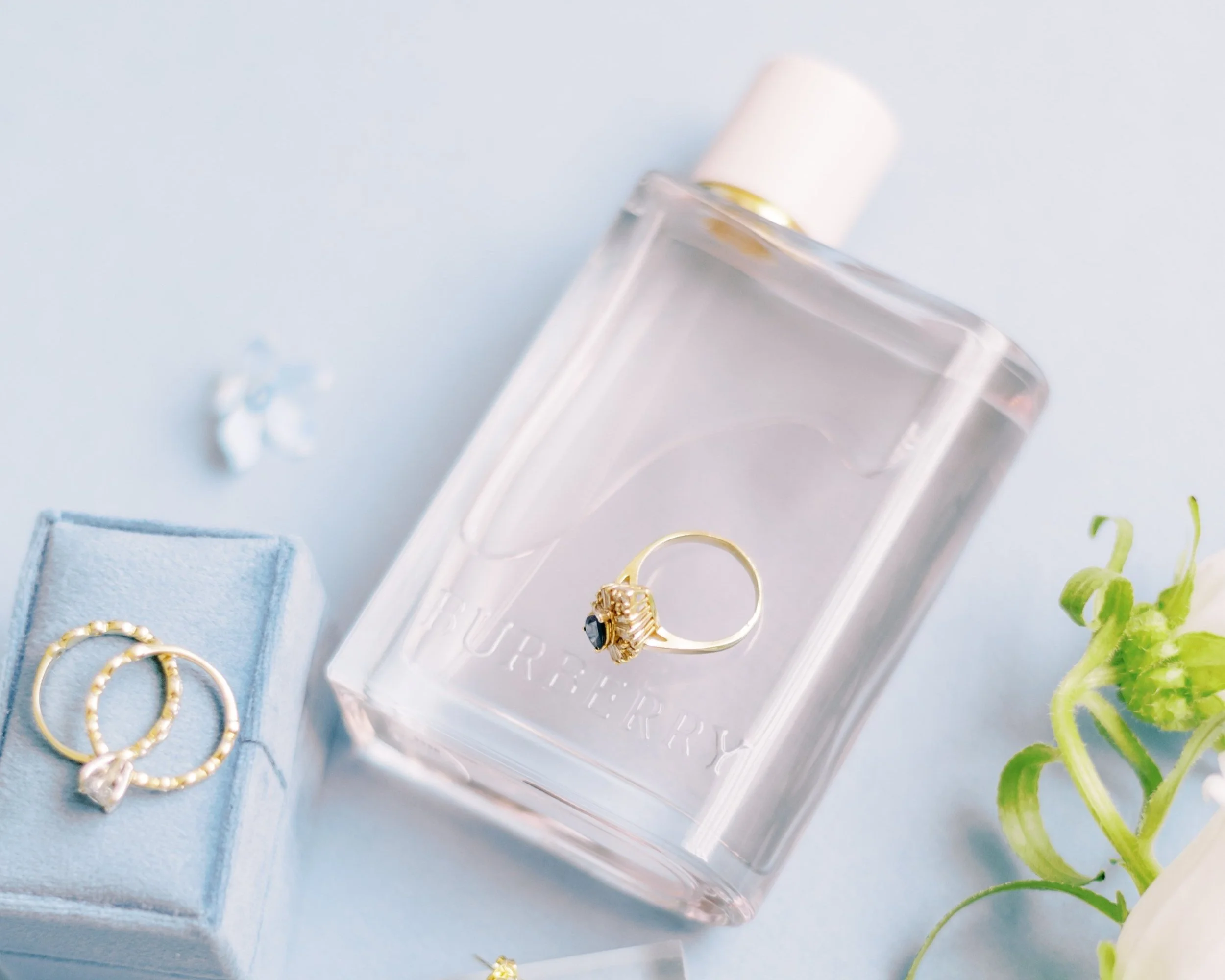 Engagement ring resting on a clear perfume bottle beside a blue ring box on a light background.