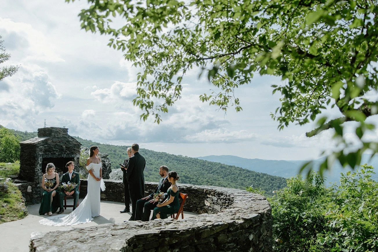 An intimate outdoor wedding ceremony taking place on a stone patio overlooking lush green mountains, showing the bride and groom at the altar with their wedding party seated nearby.