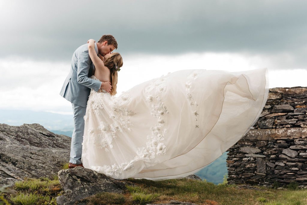 Bride and groom kissing on a windy mountain cliff with the bride’s dress flowing dramatically in the air.