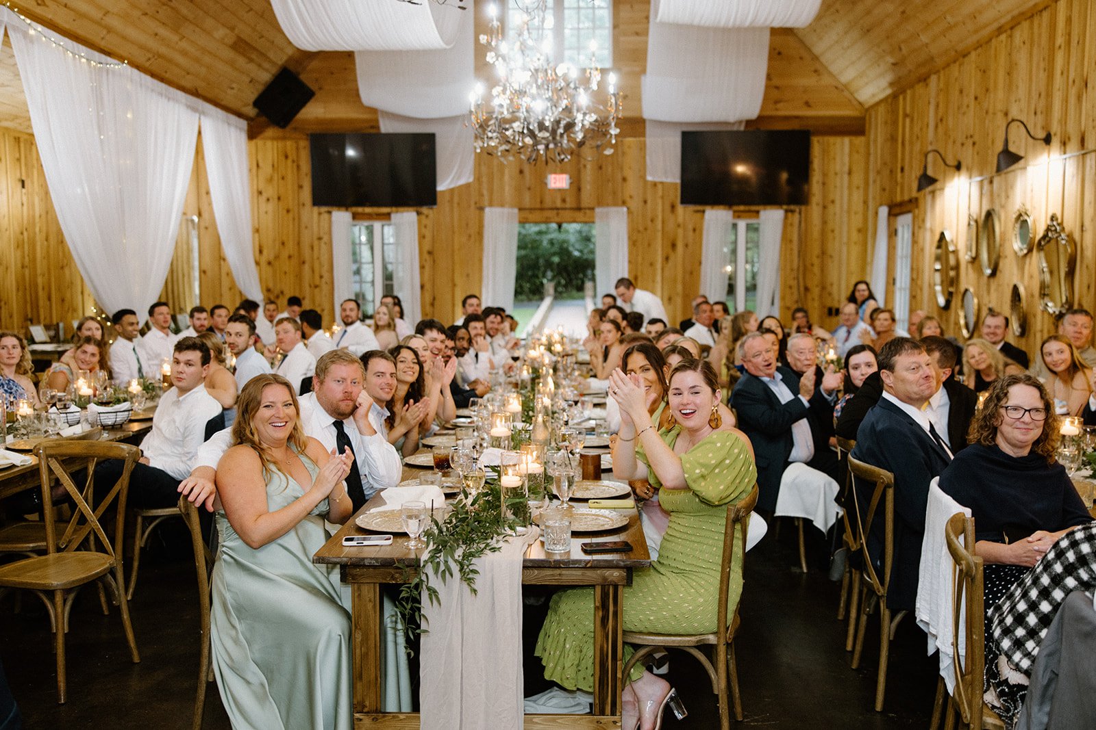 Wedding guests celebrating at long reception tables beneath chandeliers in a rustic wood reception hall.