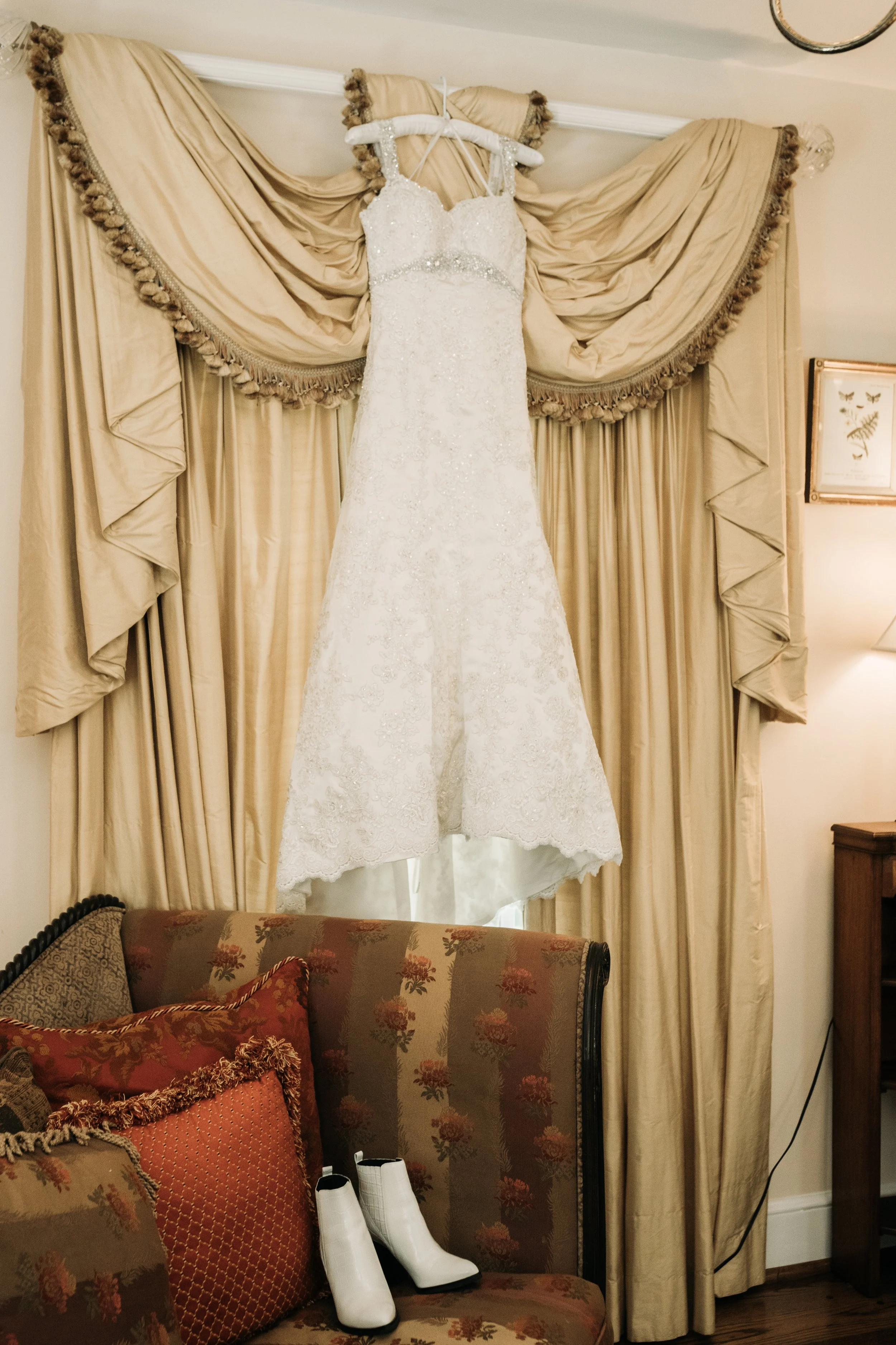 Lace wedding dress hanging above a vintage chair and white bridal boots before the ceremony.