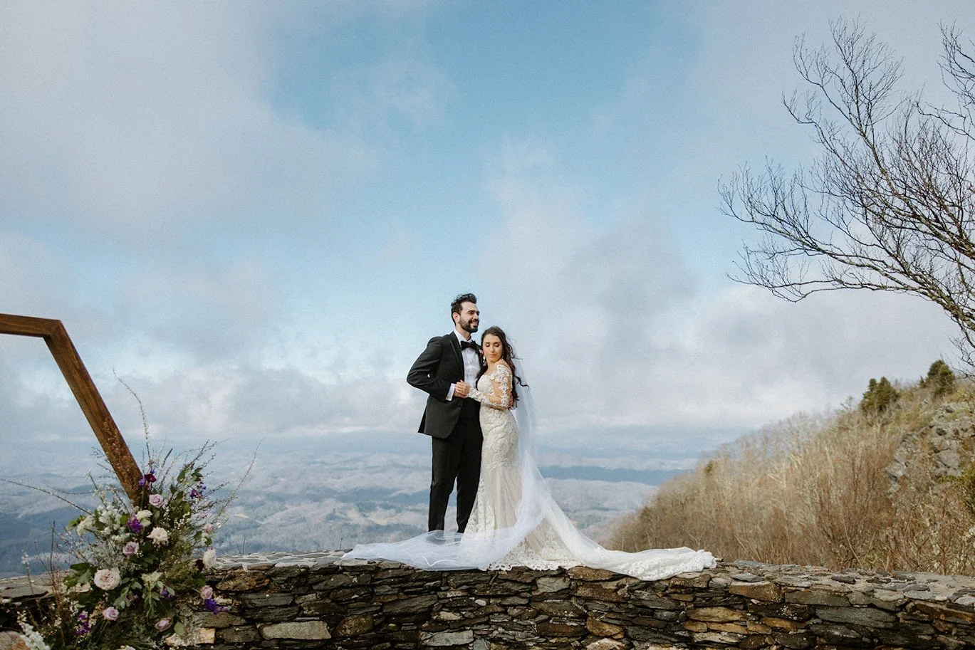 Bride and groom posing on a stone wall high above a cloudy mountain valley.