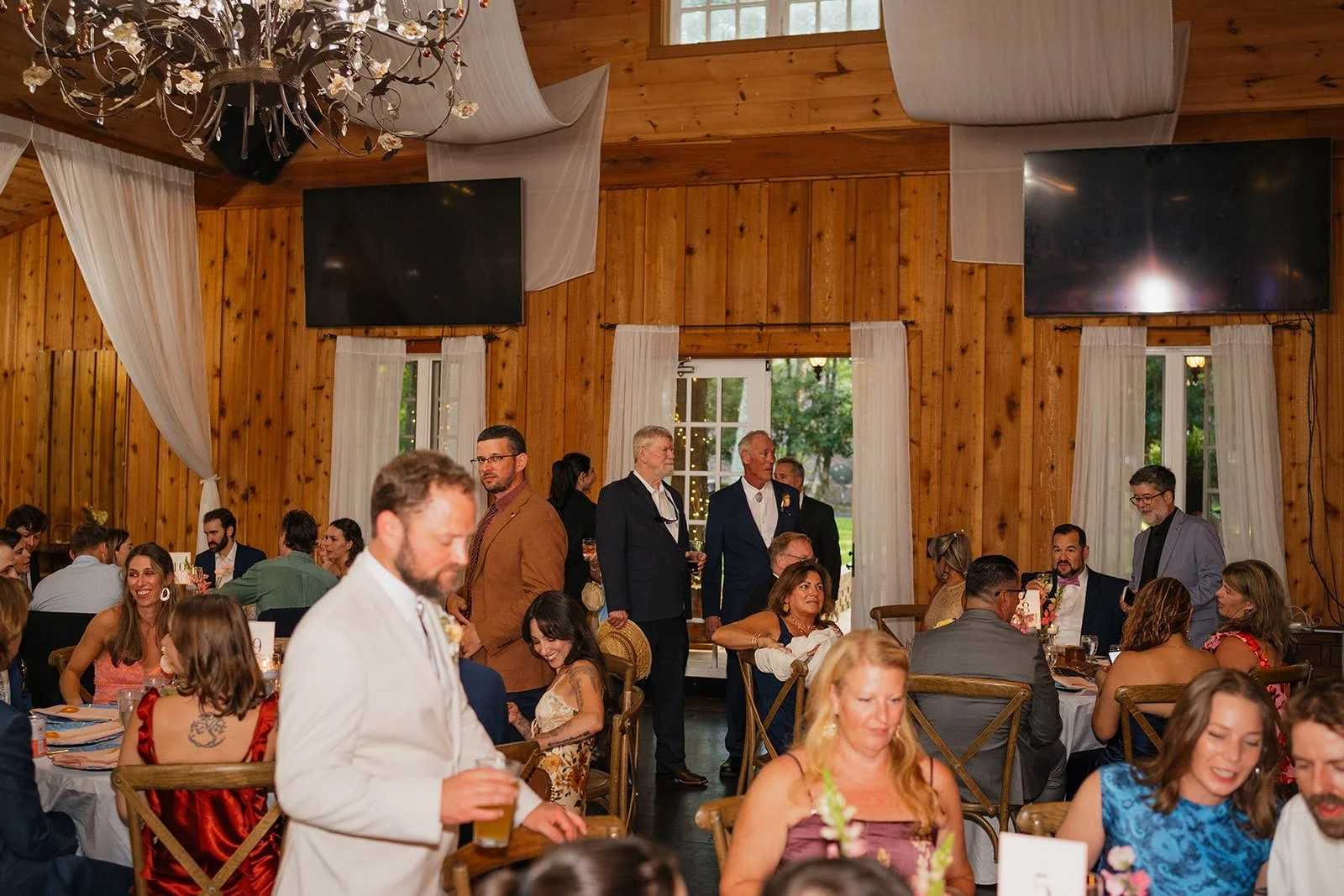 Wedding guests mingling inside a rustic wooden reception venue with chandeliers, draped fabric, and dinner tables.