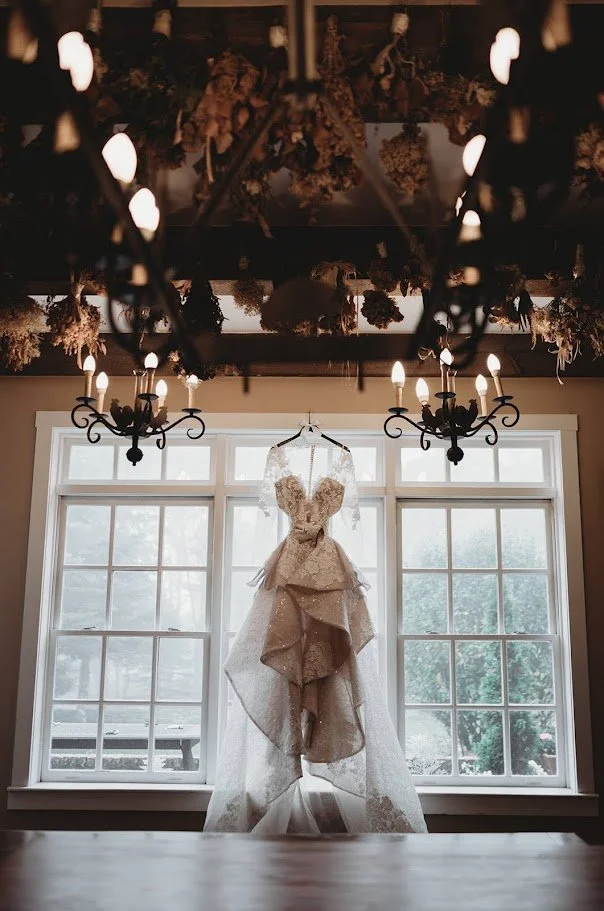 Wedding dress hanging in front of large windows under chandeliers in a moody bridal details shot.