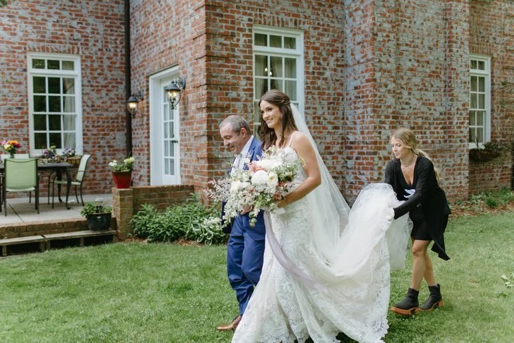 Smiling bride walks down the aisle at her wedding, holding a bouquet, with her father and a wedding planner assisting her. The wedding is taking place outdoors with a brick building in the background.