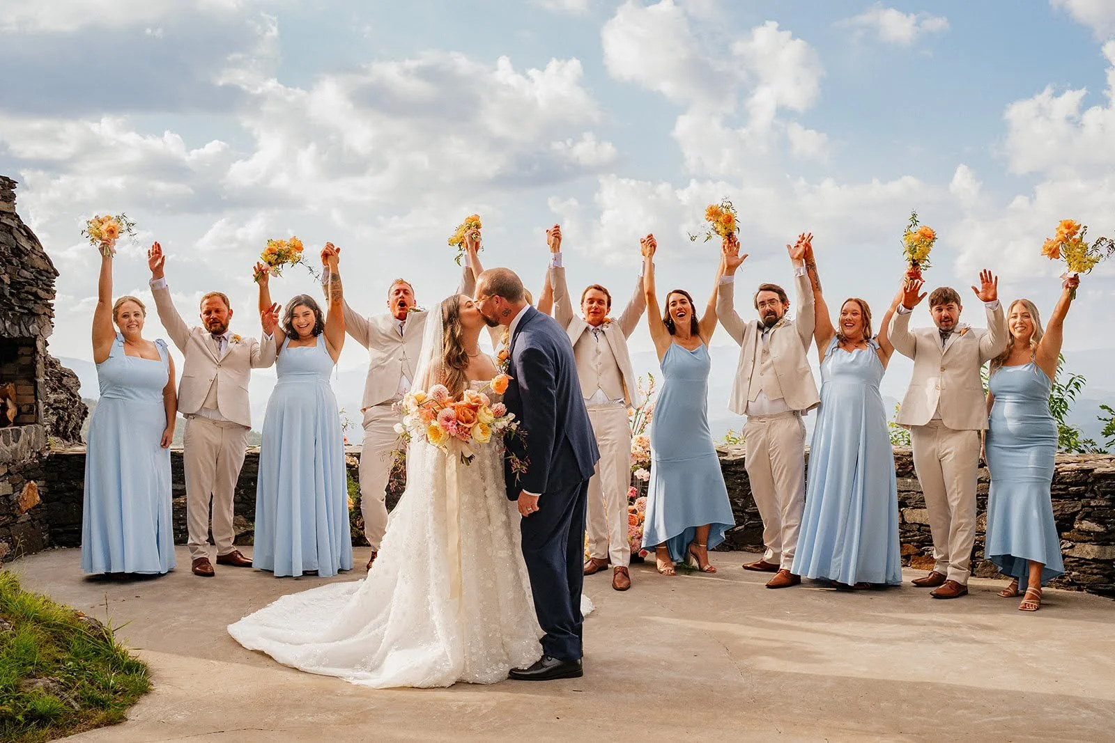 Bride and groom kissing at a mountain overlook while their wedding party cheers and raises bouquets behind them.