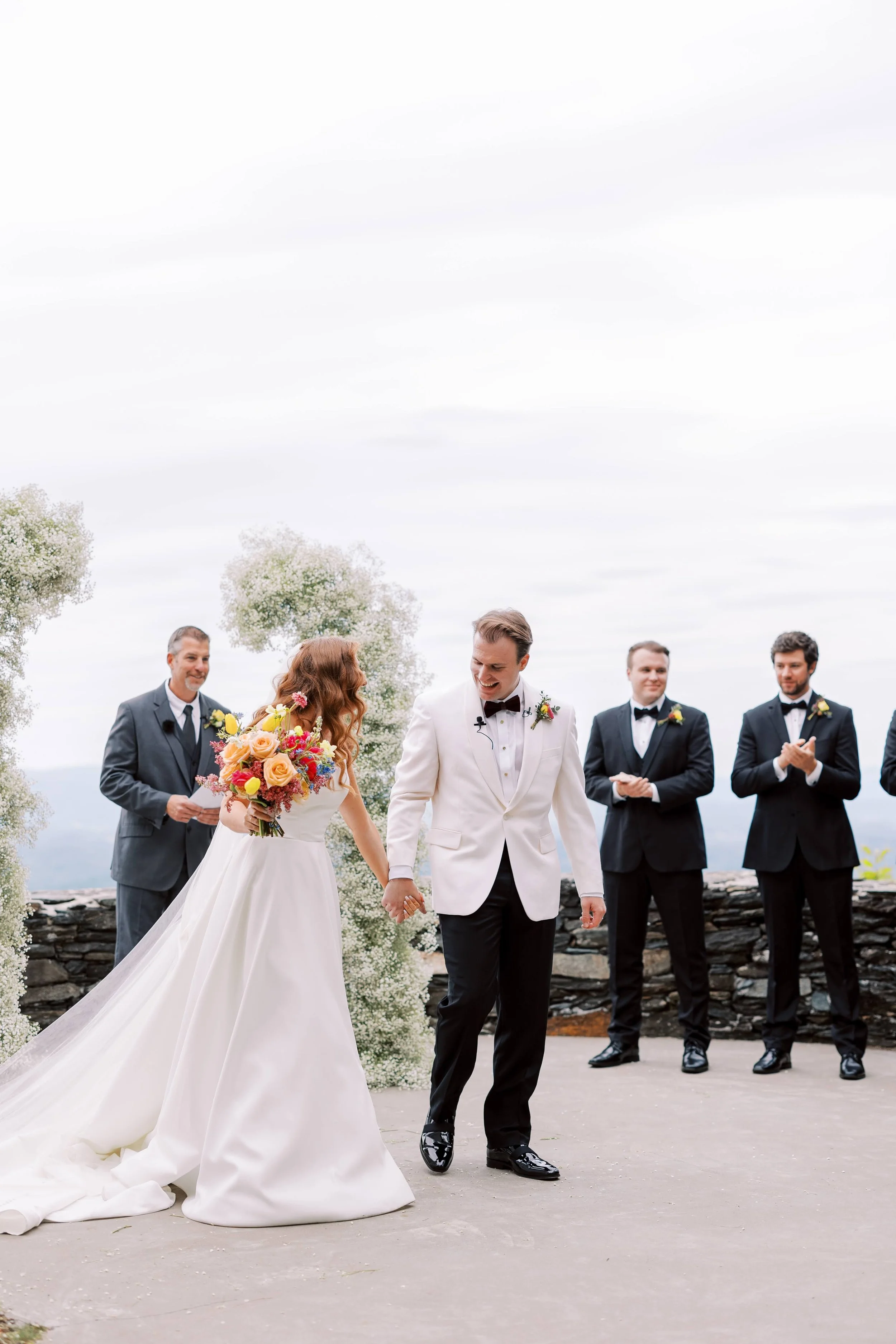 Bride and groom walking down the aisle after the ceremony, groom in a white tuxedo and bride holding a colorful bouquet.