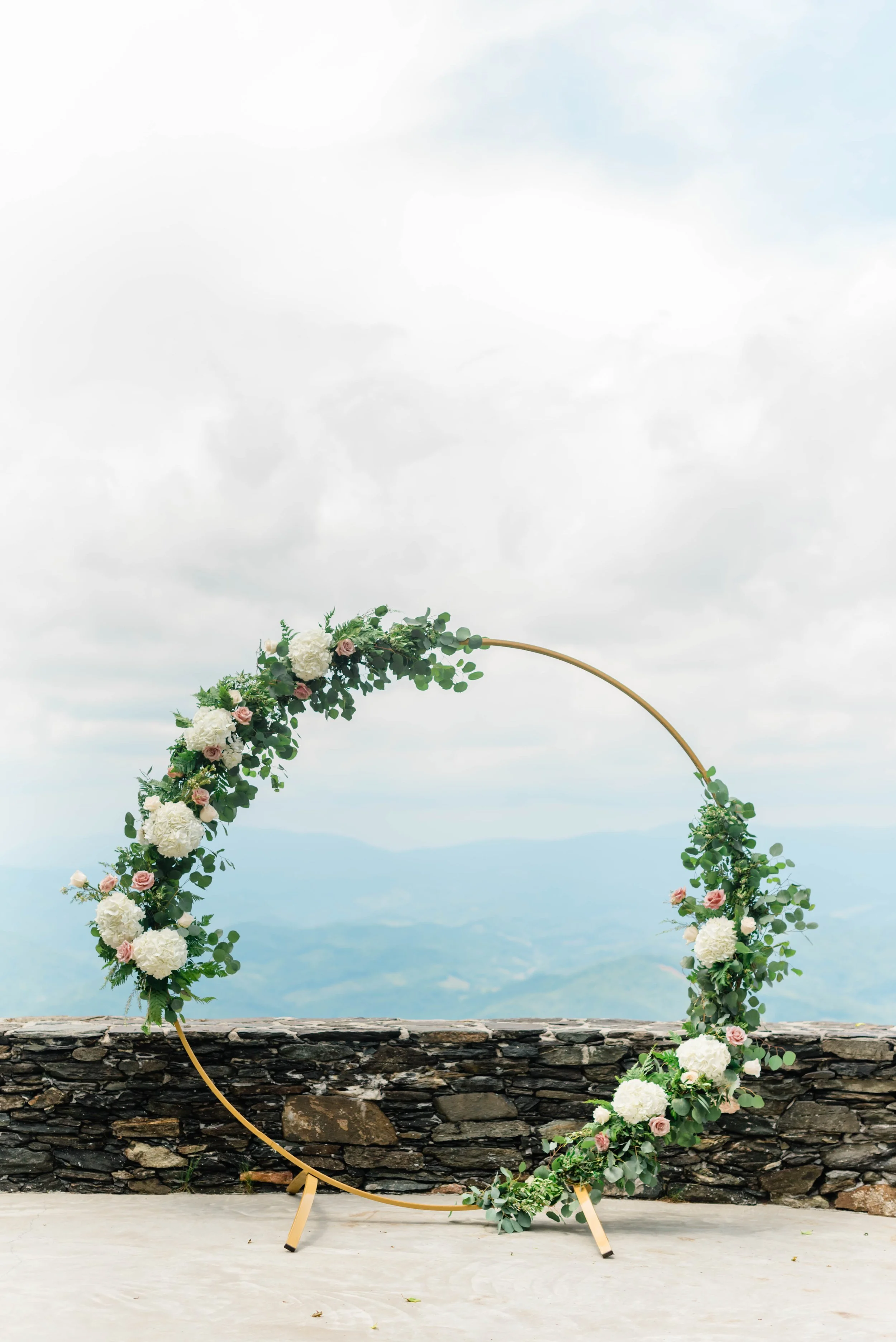 Gold circle wedding ceremony arch decorated with greenery and white flowers overlooking the mountains.
