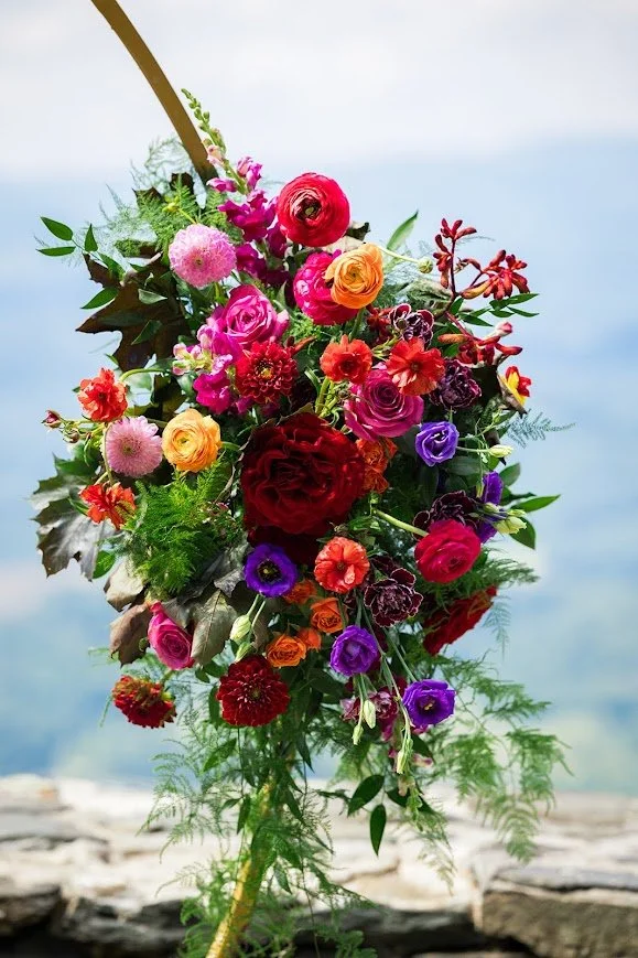 Vibrant wedding floral arrangement with roses and ranunculus in pink, red, orange, and purple on a curved arch.