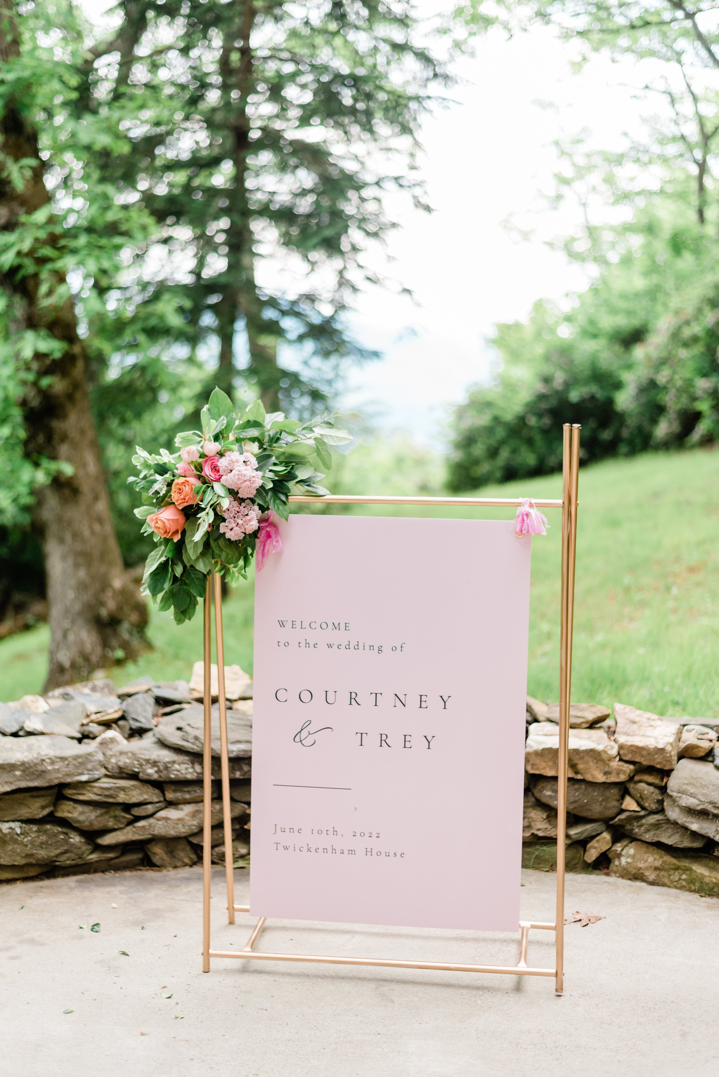Pink wedding welcome sign decorated with a colorful floral arrangement on a gold stand.