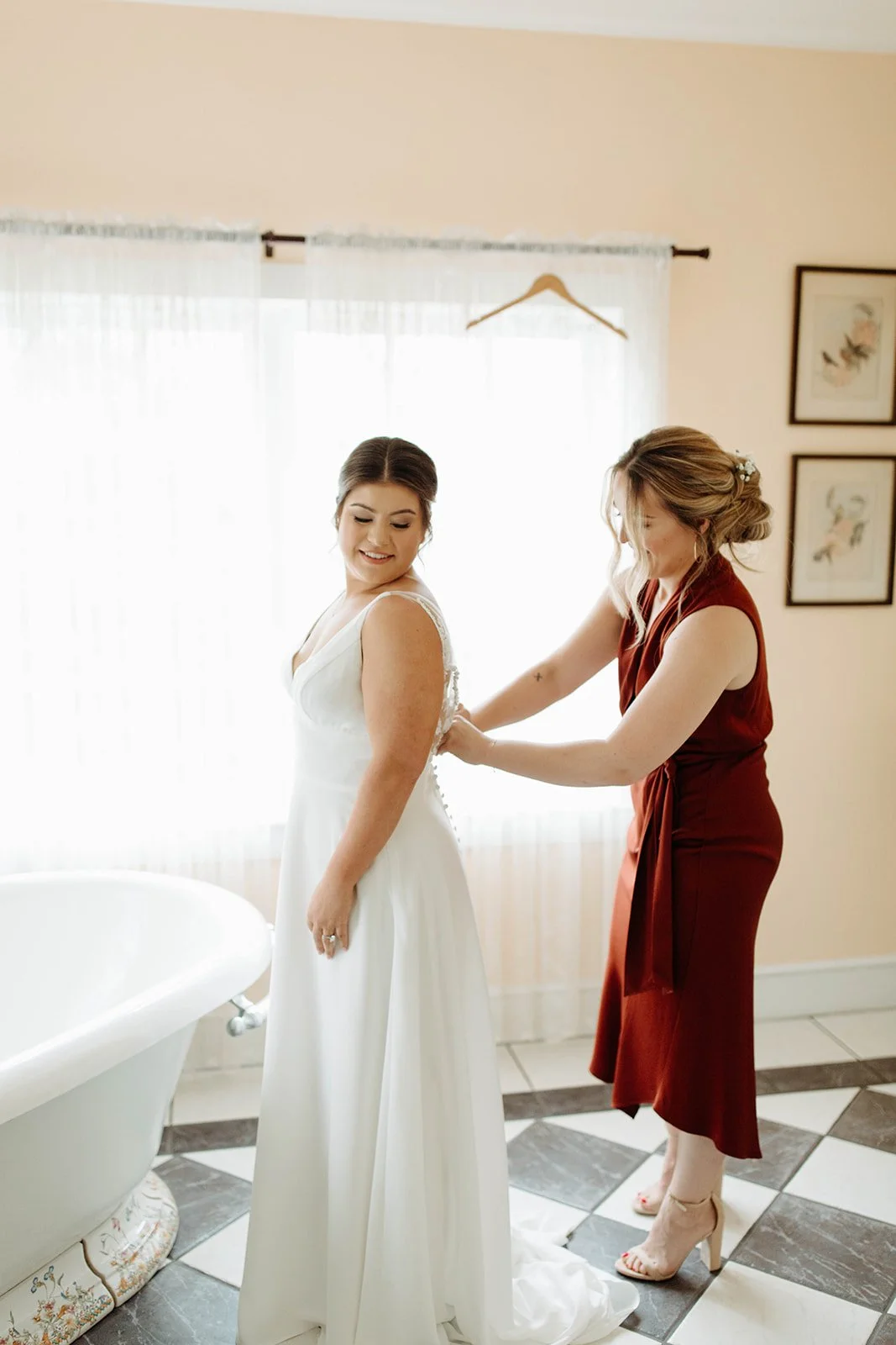 Bridesmaid helping the bride button her dress in a bright room with checkered tile floor.