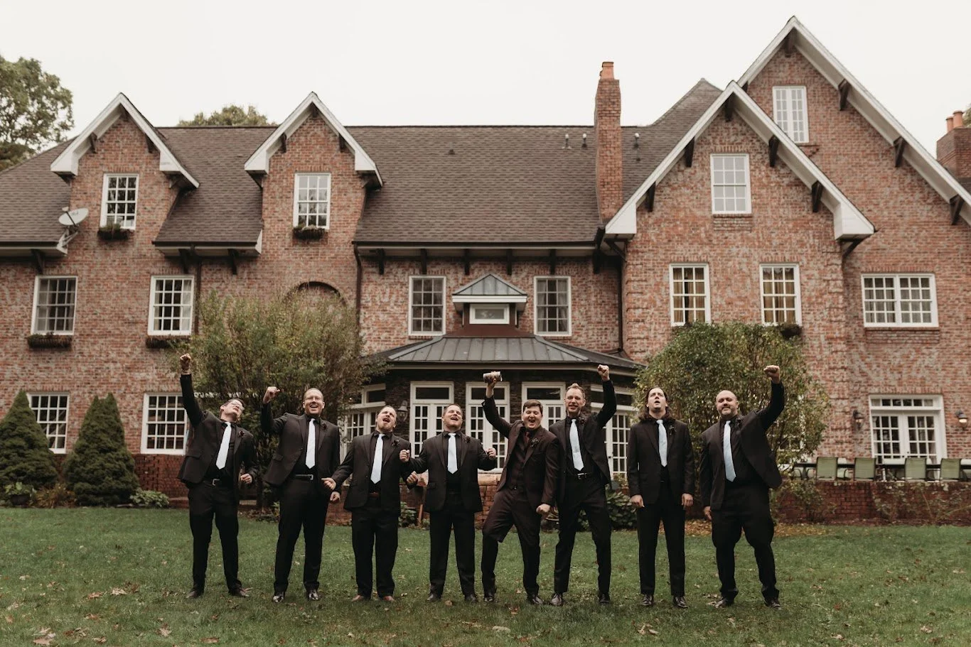 Groom and groomsmen in black suits posing outside a historic brick estate wedding venue.