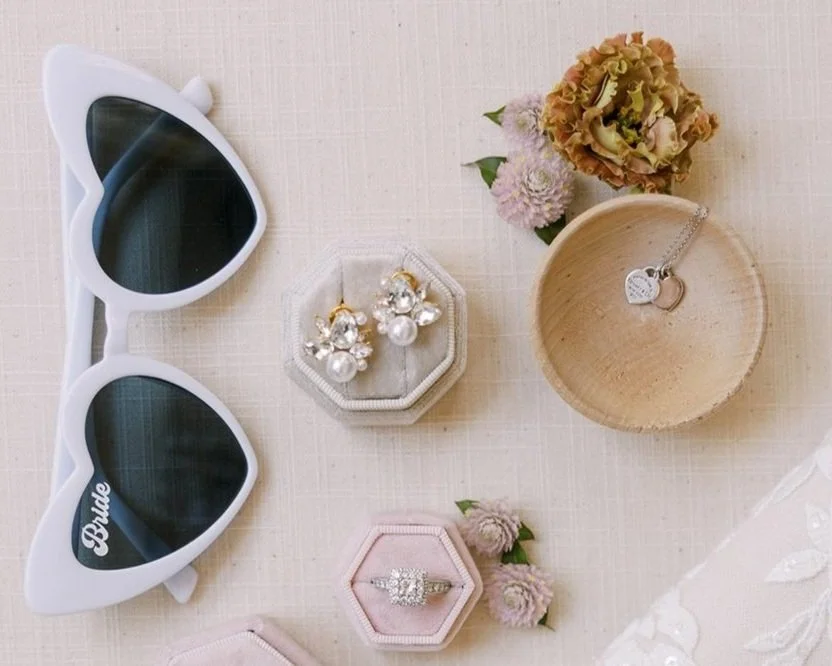 Flat lay of white sunglasses, ring boxes, earrings, and engagement ring on a neutral fabric background.