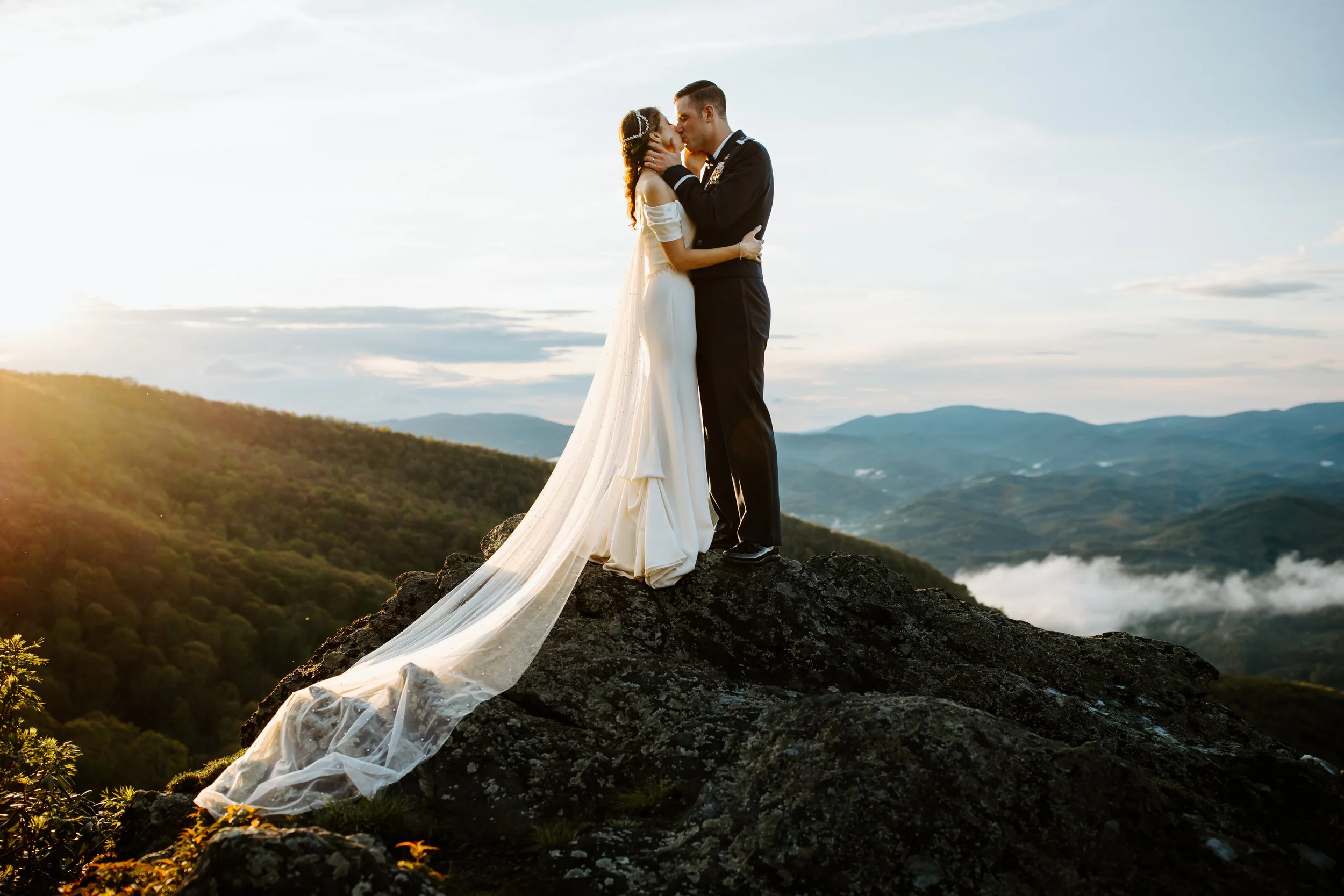 Bride and groom kissing at sunset on a rocky overlook with mountains in the background.