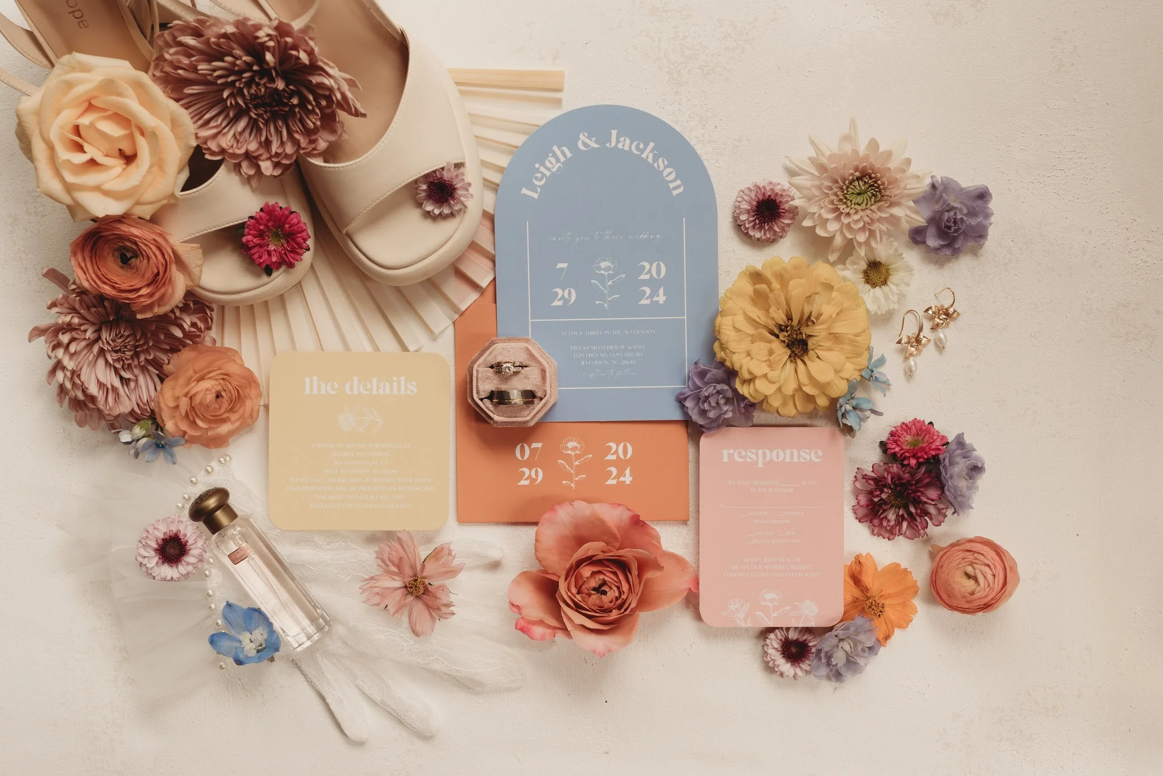 Wedding details flatlay with invitation suite, shoes, and pastel flowers arranged on a light background.