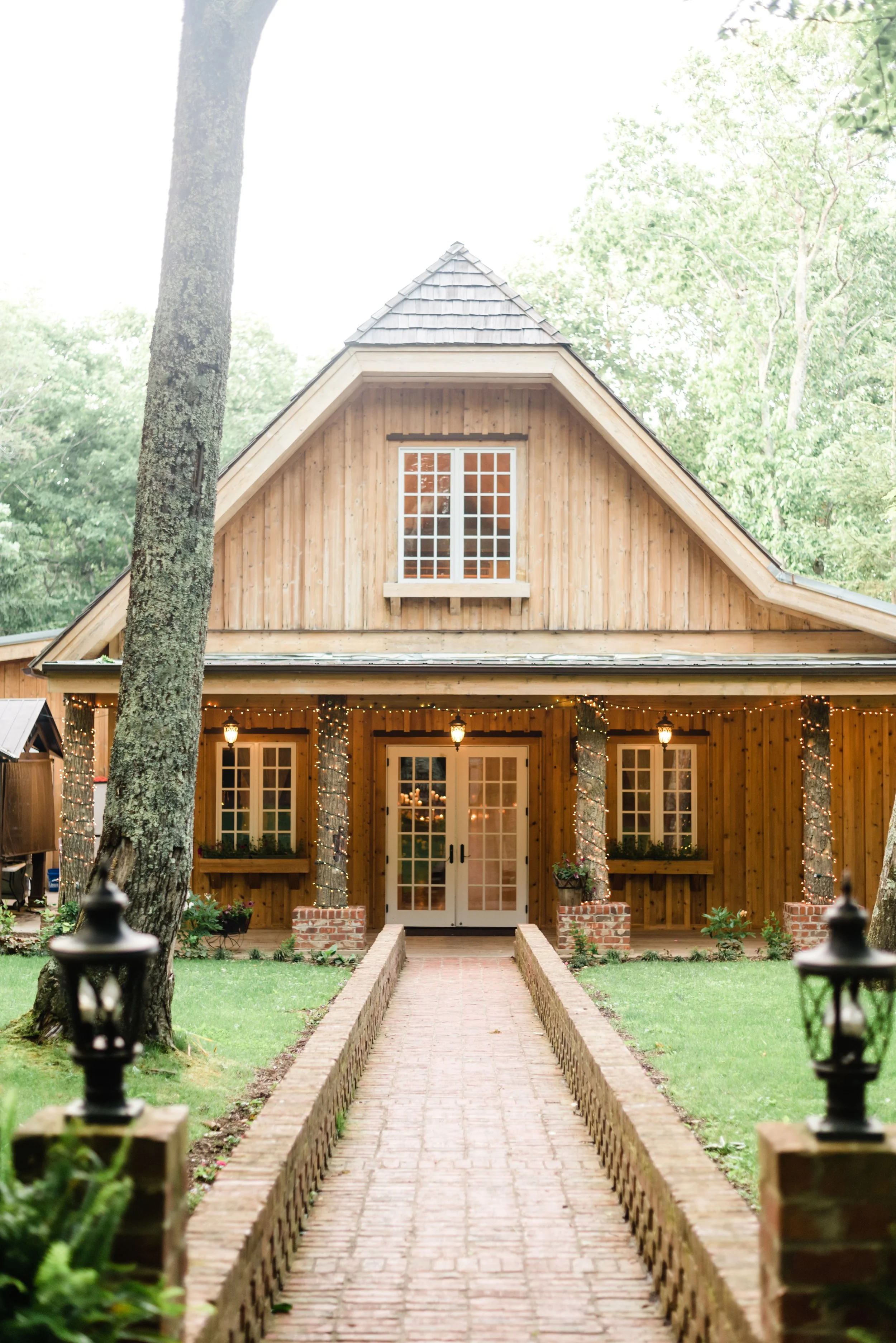 Rustic wood wedding chapel exterior with French doors, brick walkway, trees, and warm string lights.