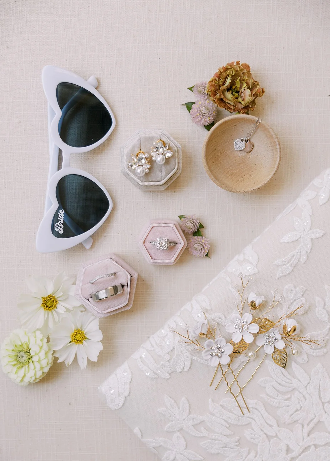 Flat lay of white sunglasses, ring boxes, earrings, and engagement ring on a neutral fabric background.