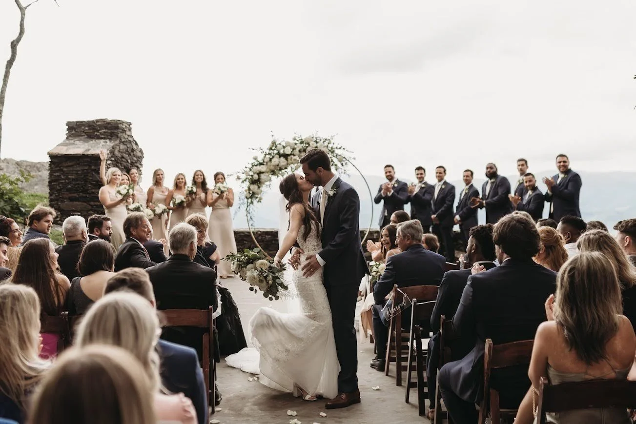 Wedding couple sharing their first kiss at a mountain ceremony as guests cheer.