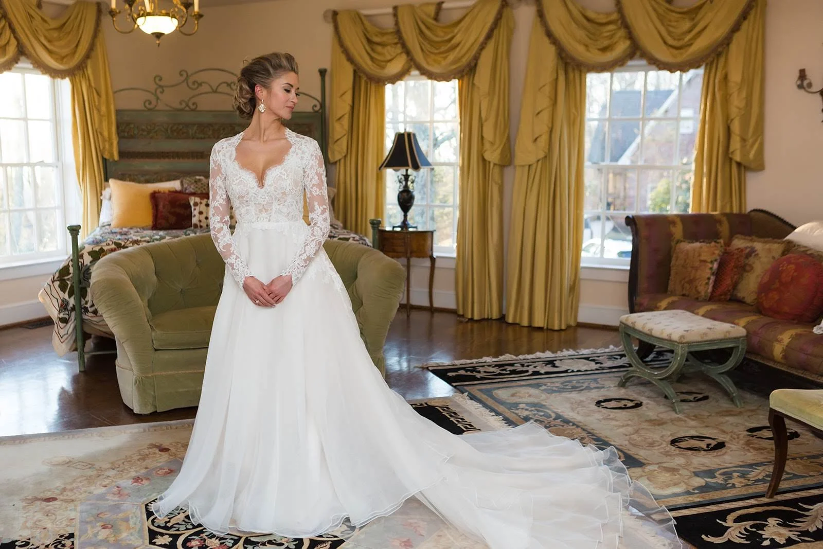 Bride standing indoors in a lace-sleeve wedding dress with a long train.
