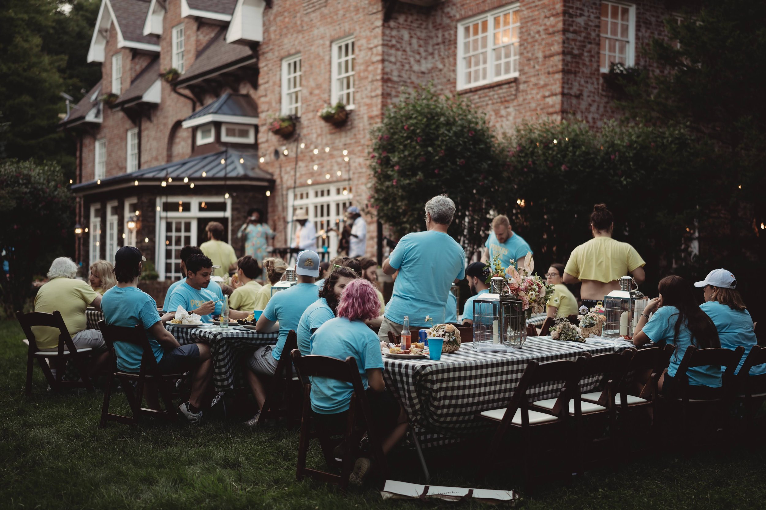 Outdoor rehearsal dinner on the lawn with long tables and guests seated in front of a brick estate