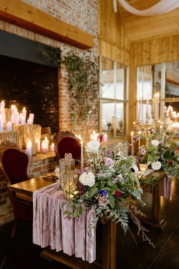 Sweetheart table with lush florals and candles set in front of a brick fireplace at a rustic wedding reception