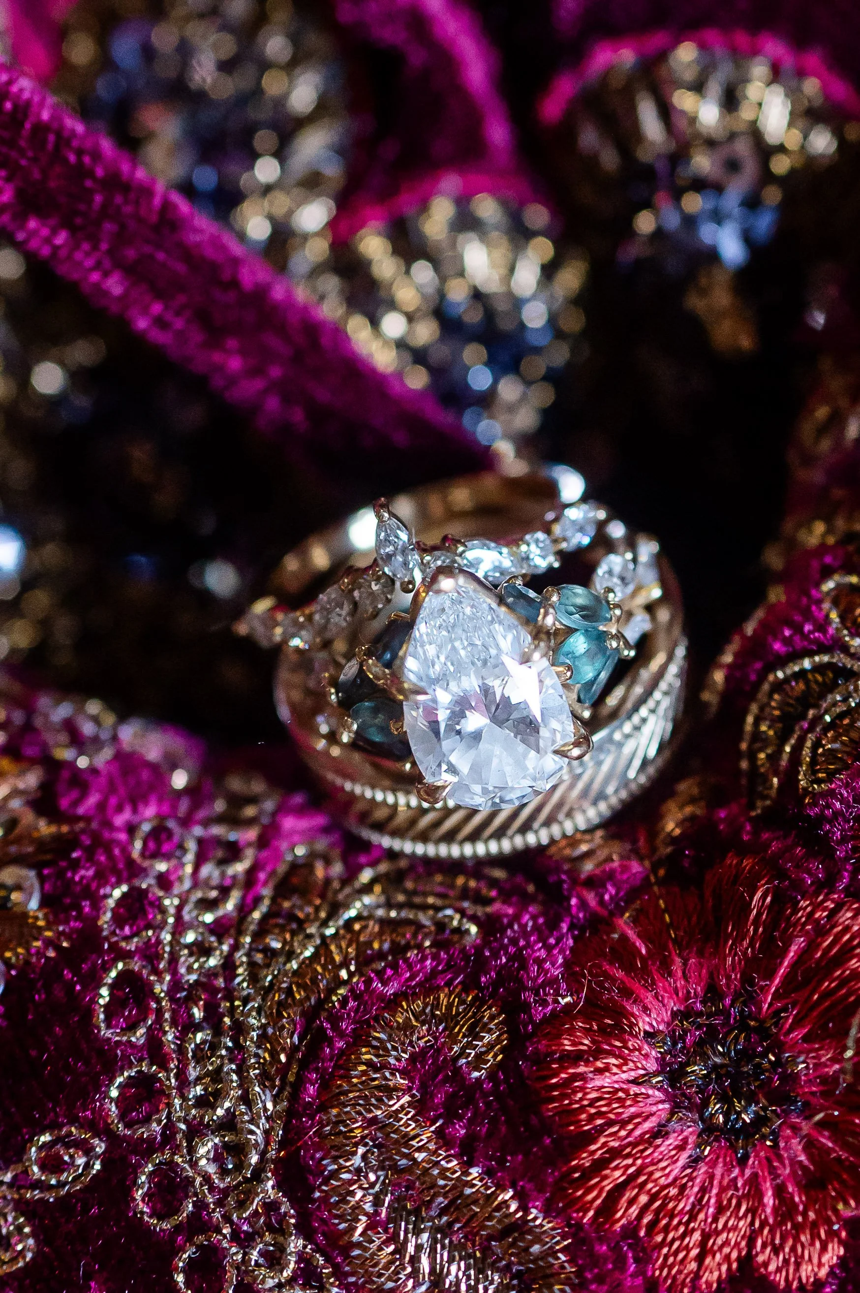 Pear-shaped engagement ring photographed on vibrant embroidered fabric with gold and magenta details.