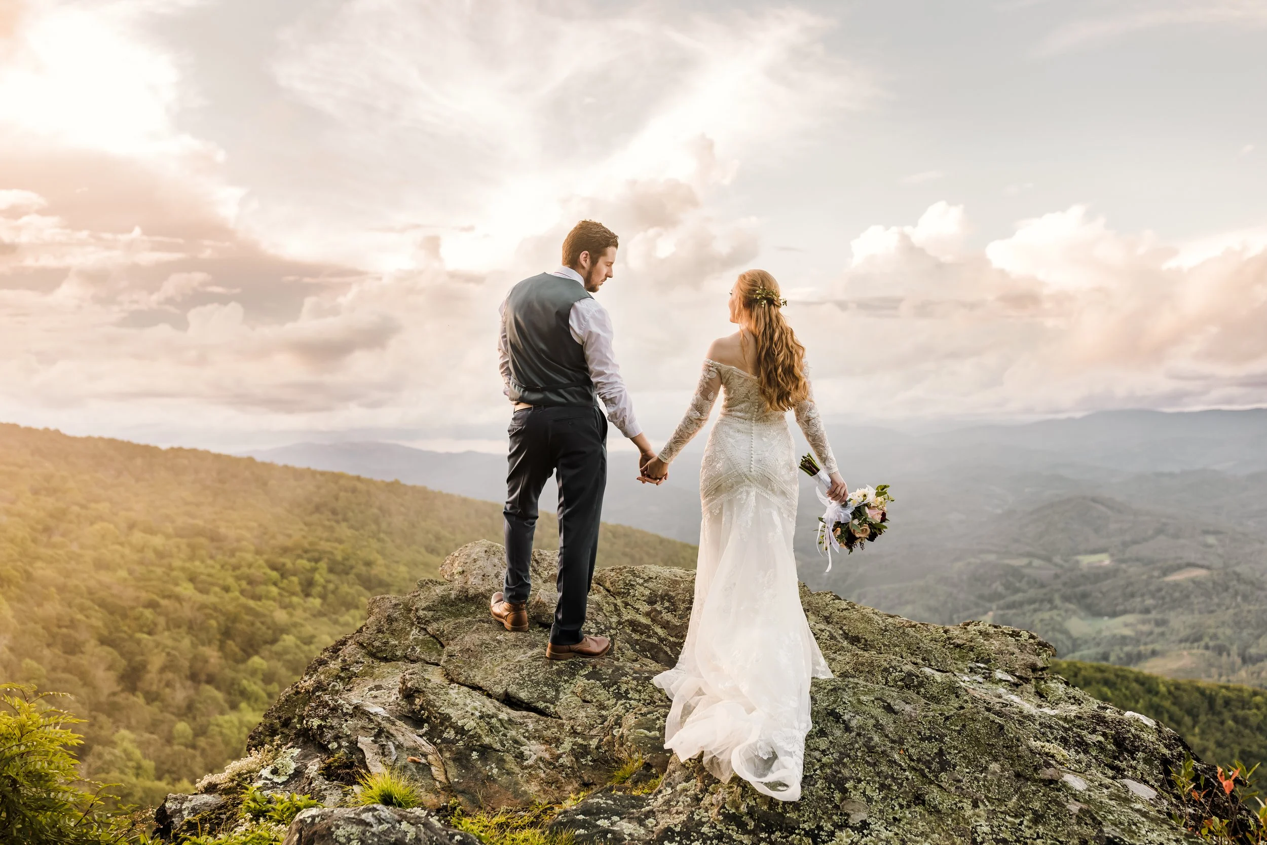 Bride and groom holding hands on a rocky mountain cliff at sunset with layered mountain views