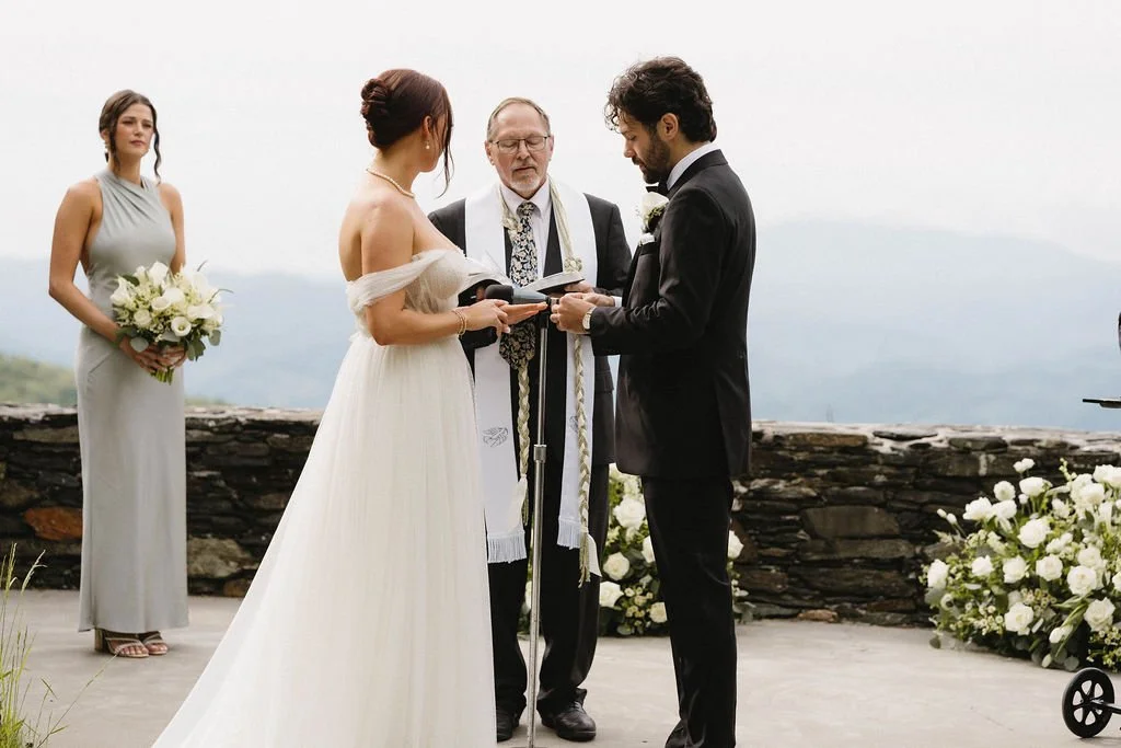 Wedding guests seated in wooden chairs during an outdoor mountain ceremony surrounded by trees and stone walls.