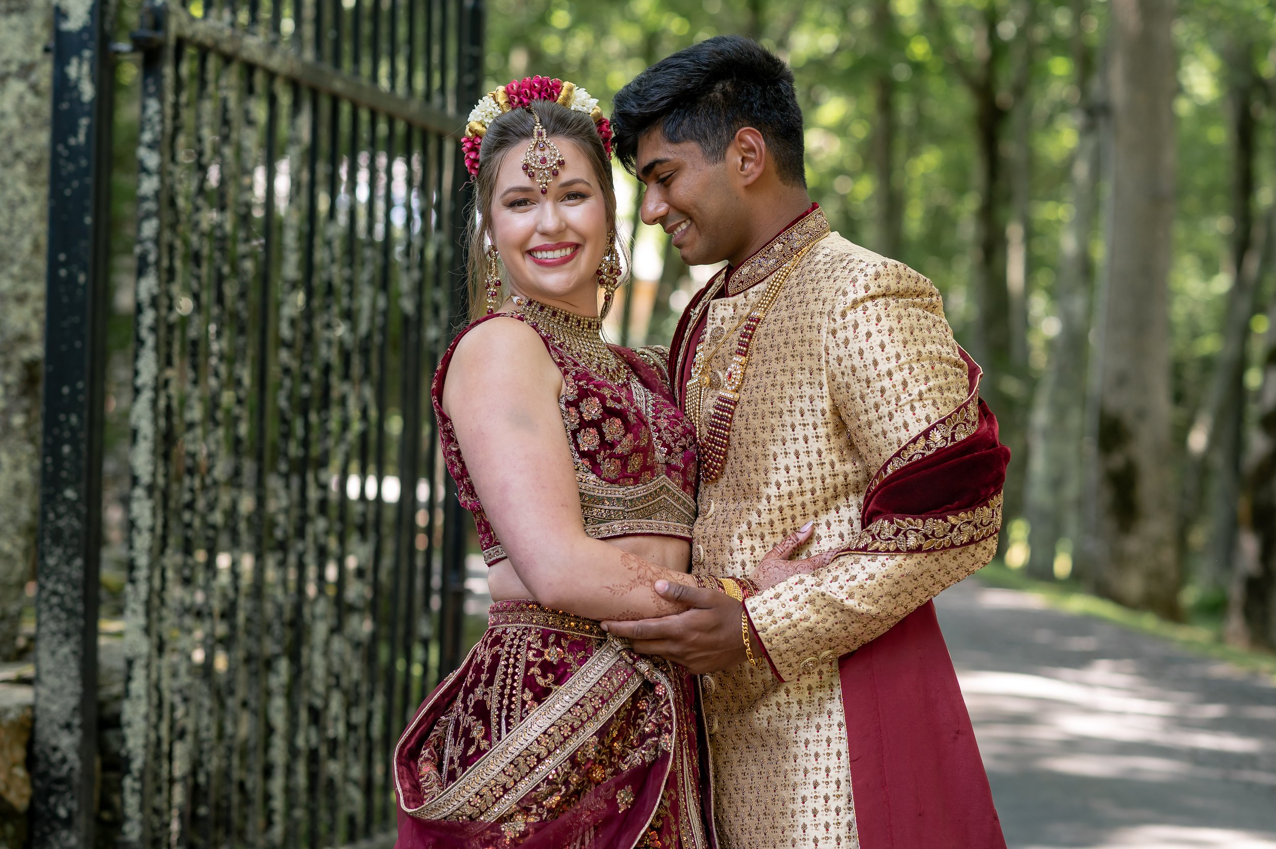 South Asian wedding couple smiling together in red and gold traditional attire beside an iron gate.