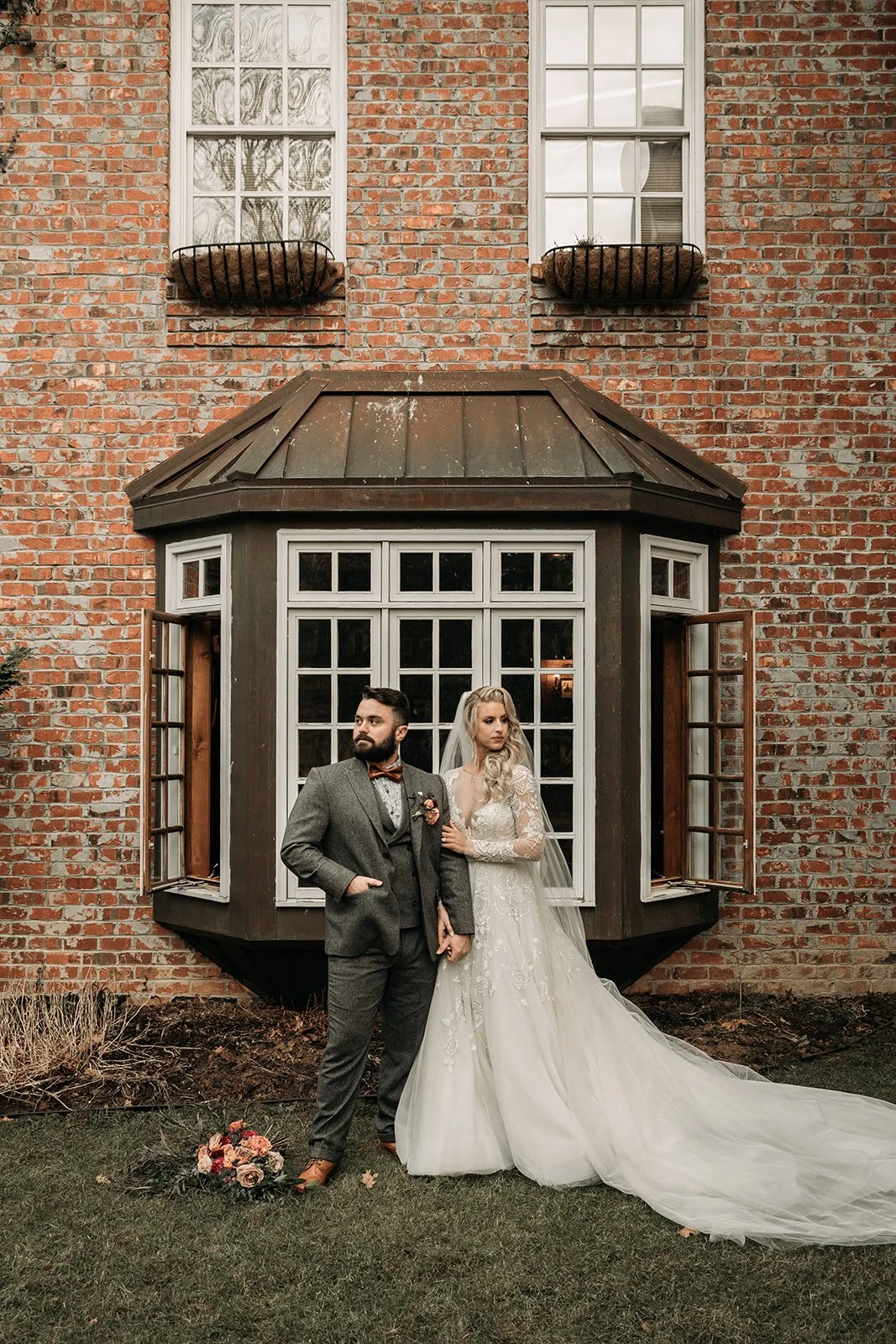 Bride and groom standing hand in hand outside a brick wedding venue with a large bay window.