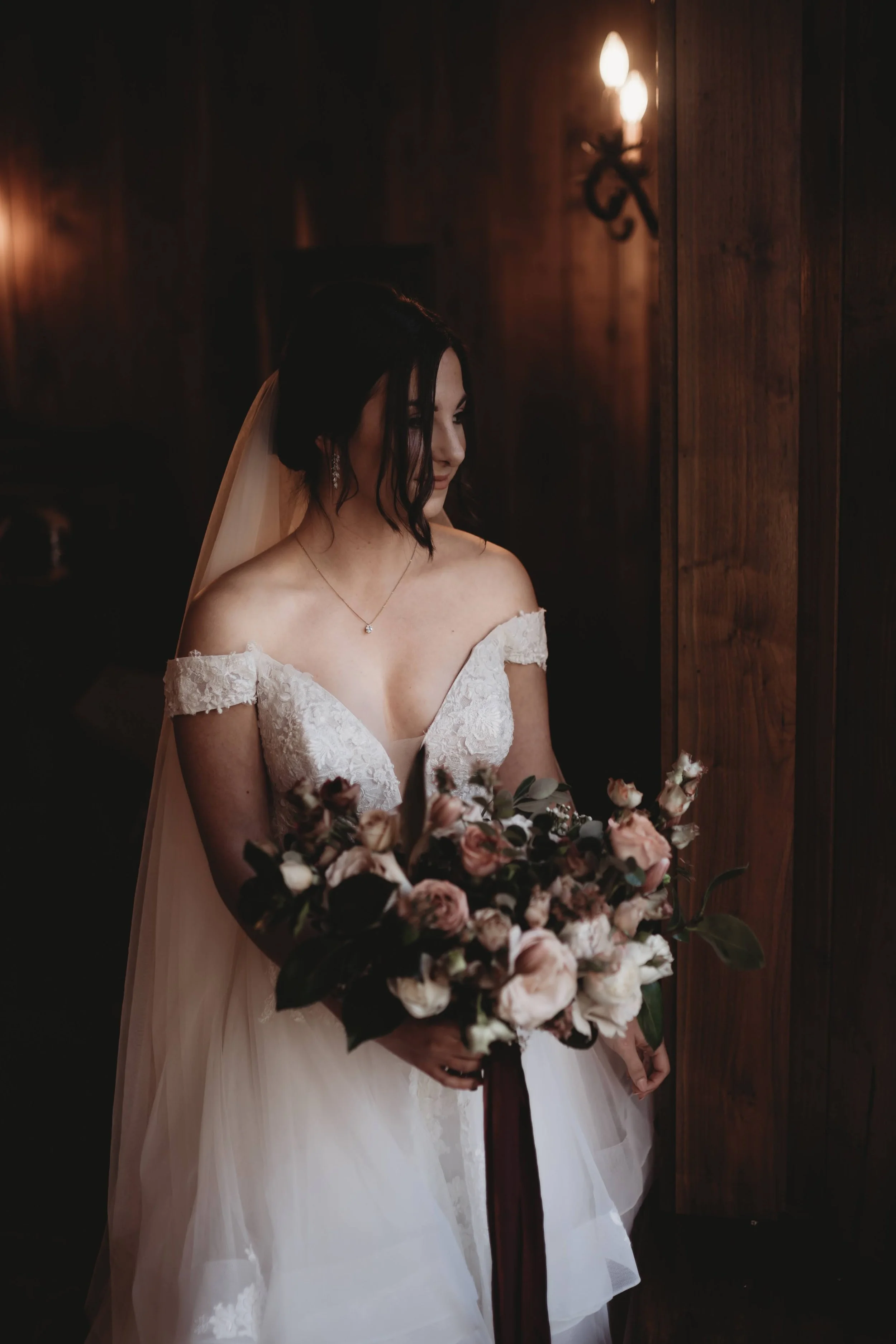 Bride in an off-the-shoulder wedding gown holding a bouquet in warm indoor window light.