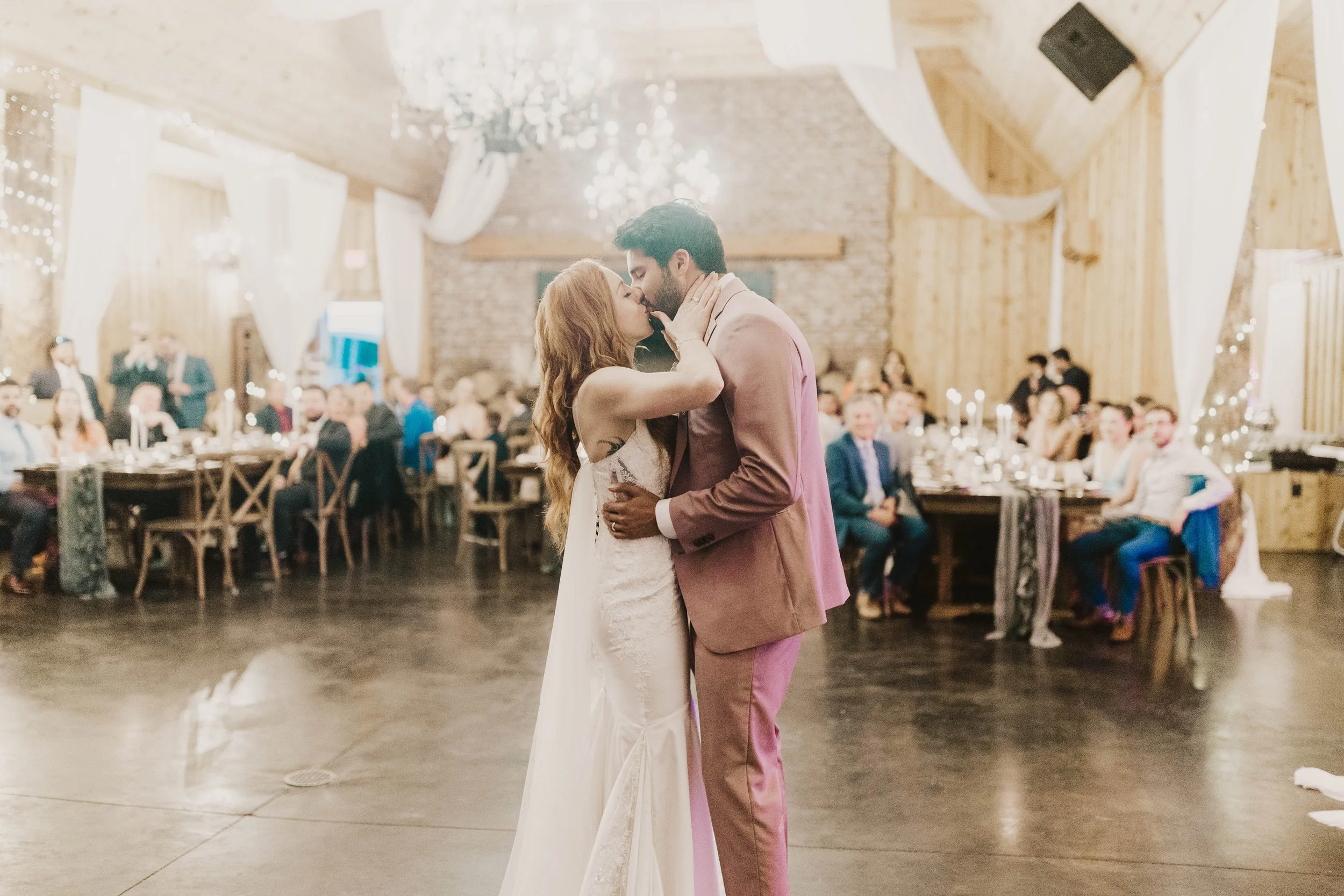 Bride and groom sharing their first dance in a rustic indoor reception hall with seated guests, draped fabric, and soft lighting.