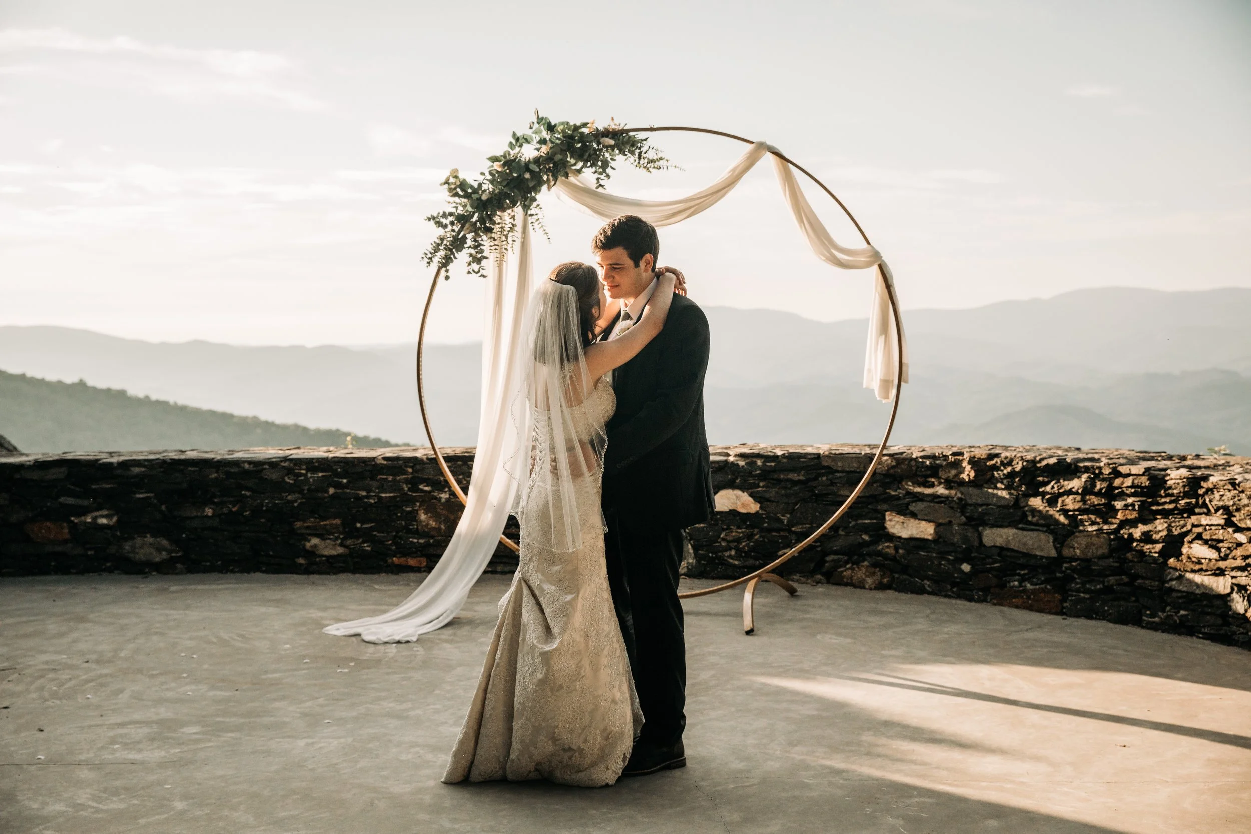 Bride and groom kissing beneath a circular wedding arch with white draping and mountain views behind them.Bride and groom kissing beneath a circular wedding arch with white draping and mountain views behind them.