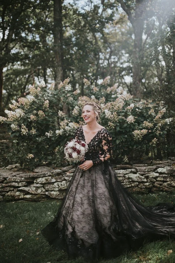 Bride smiling with a bouquet while wearing a black lace wedding dress near a stone wall and flowering trees.