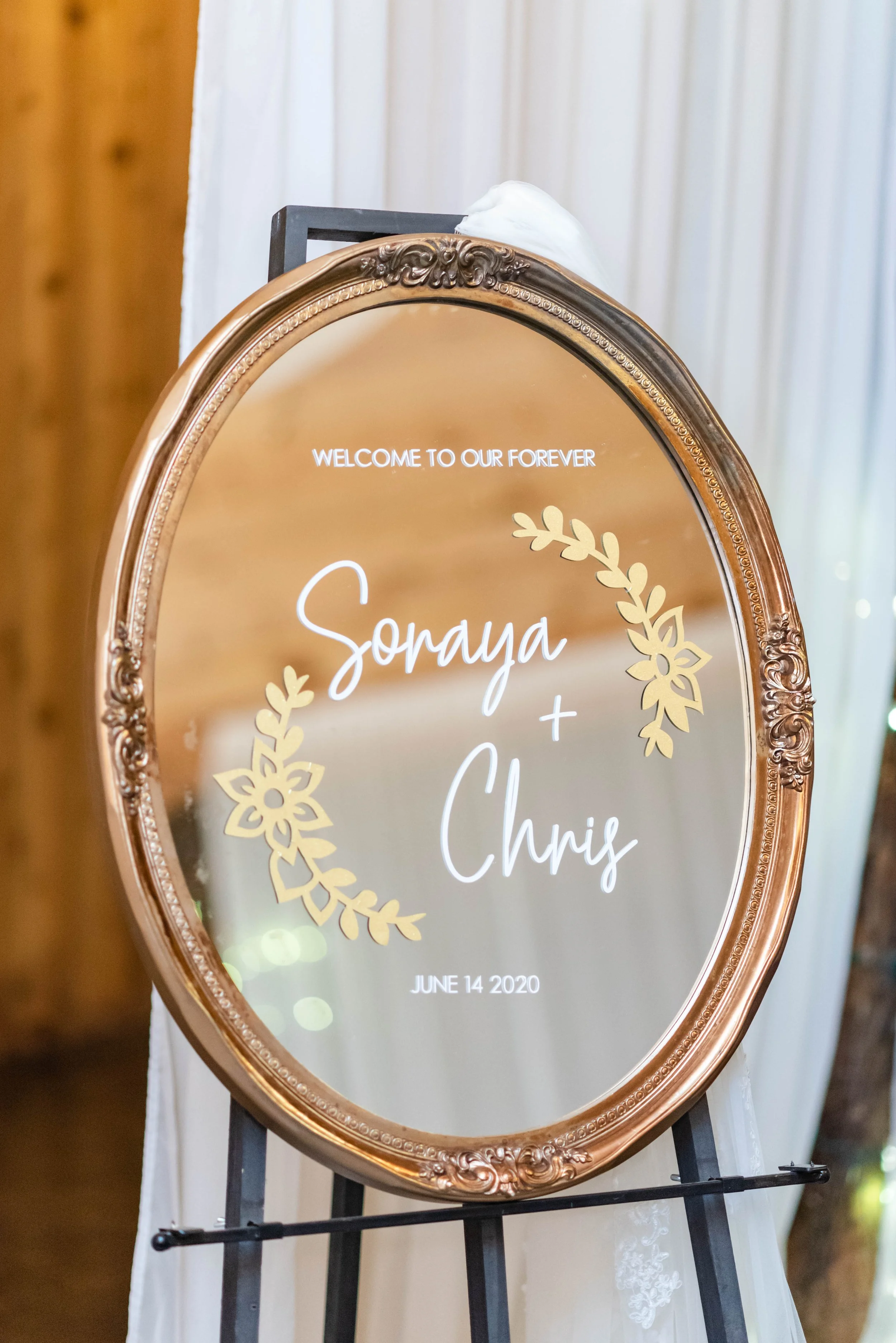 Oval mirror wedding welcome sign for Sonaya and Chris displayed on an easel with white draping.