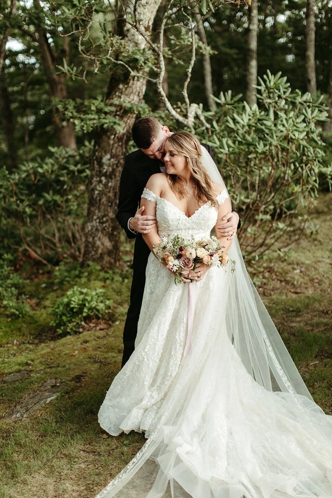 Bride and groom standing on a mountain cliff with the bride’s long veil flowing and a bouquet in hand.
