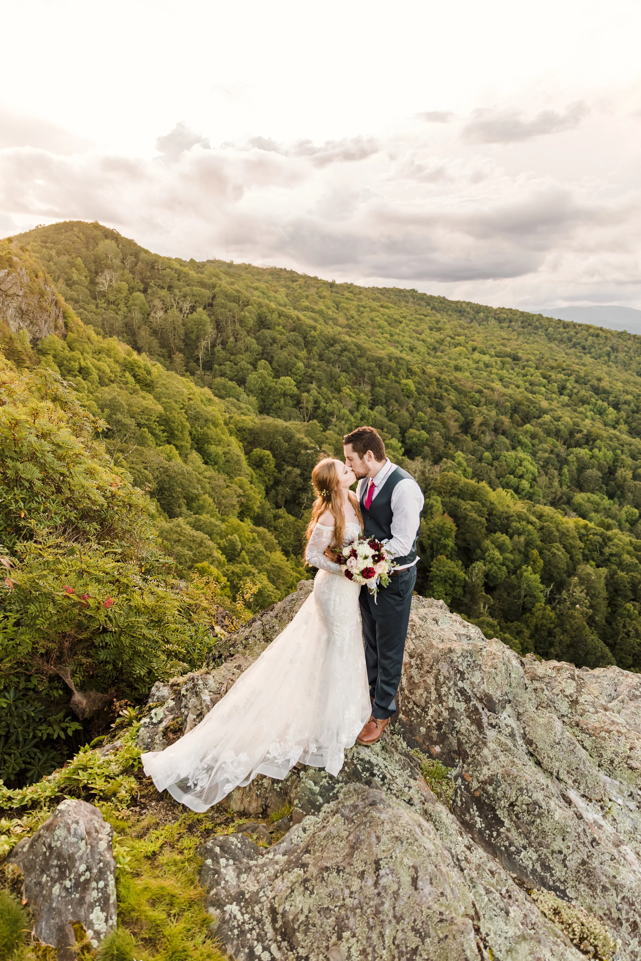 Bride and groom kissing on a cliff edge with a sweeping mountain view in the background.