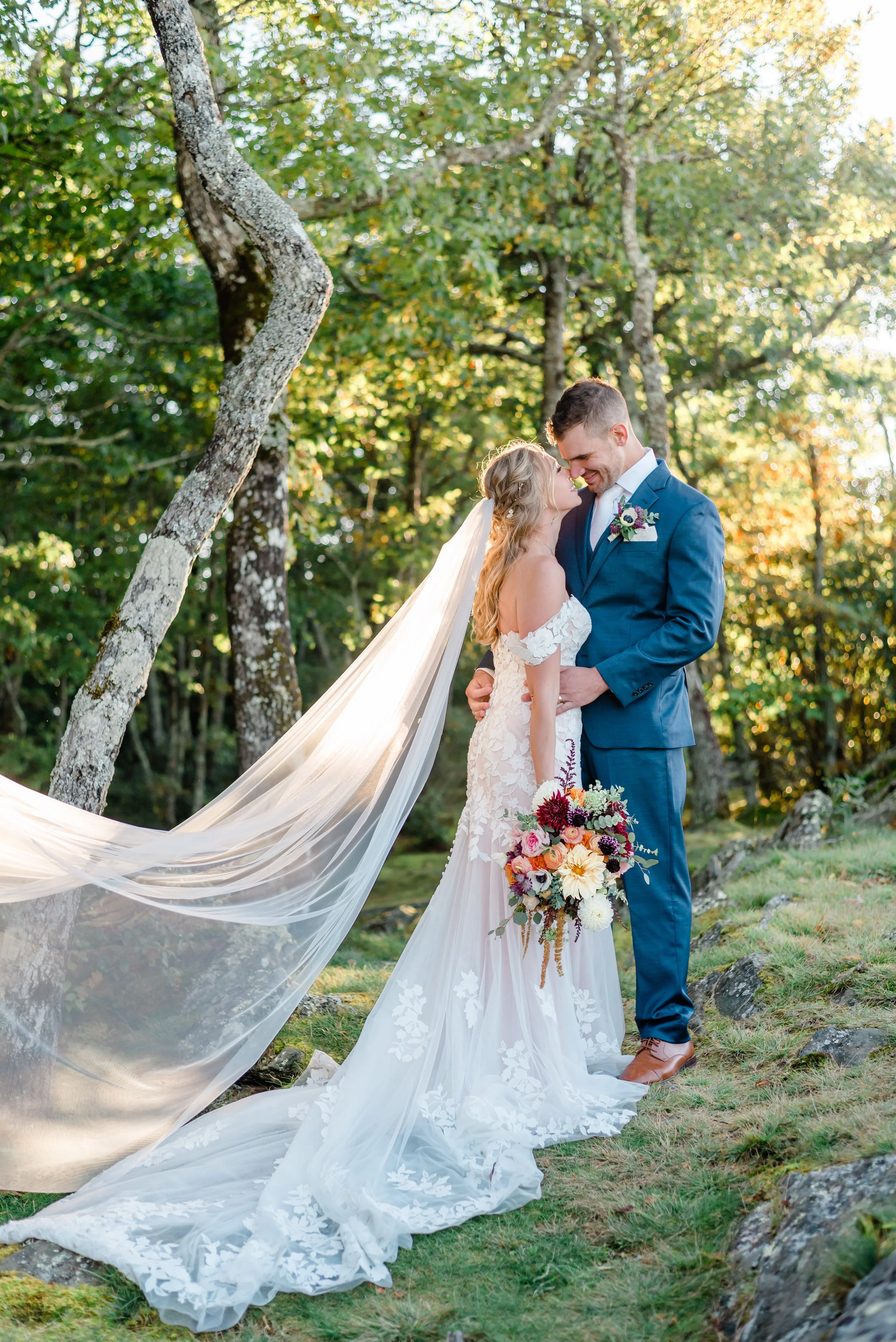 Bride and groom embracing for a romantic woodland wedding portrait with the bride’s long veil flowing behind her.