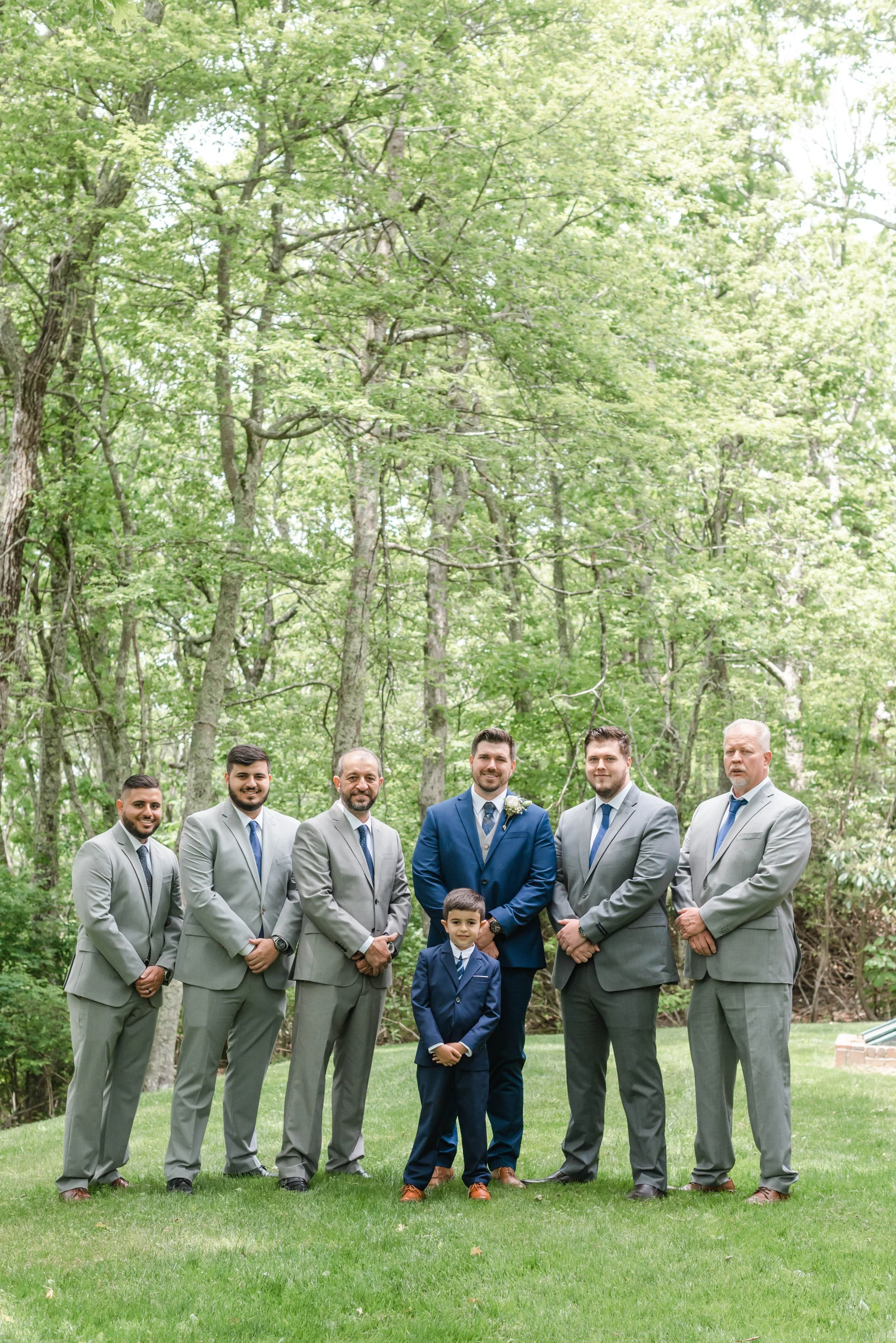 Groom and groomsmen in gray suits posing outdoors with a young ring bearer in a blue suit.