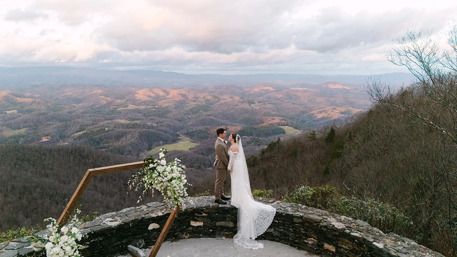 Bride and groom kissing at a scenic mountain ceremony overlook with a wooden floral arch and expansive valley views.