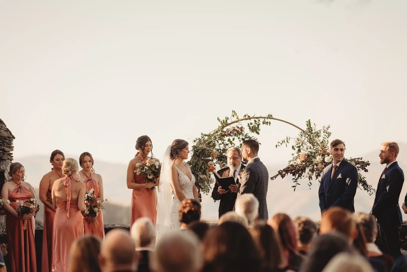 Outdoor mountain wedding ceremony with a circular greenery arch, bridesmaids in terracotta dresses, and groomsmen in suits
