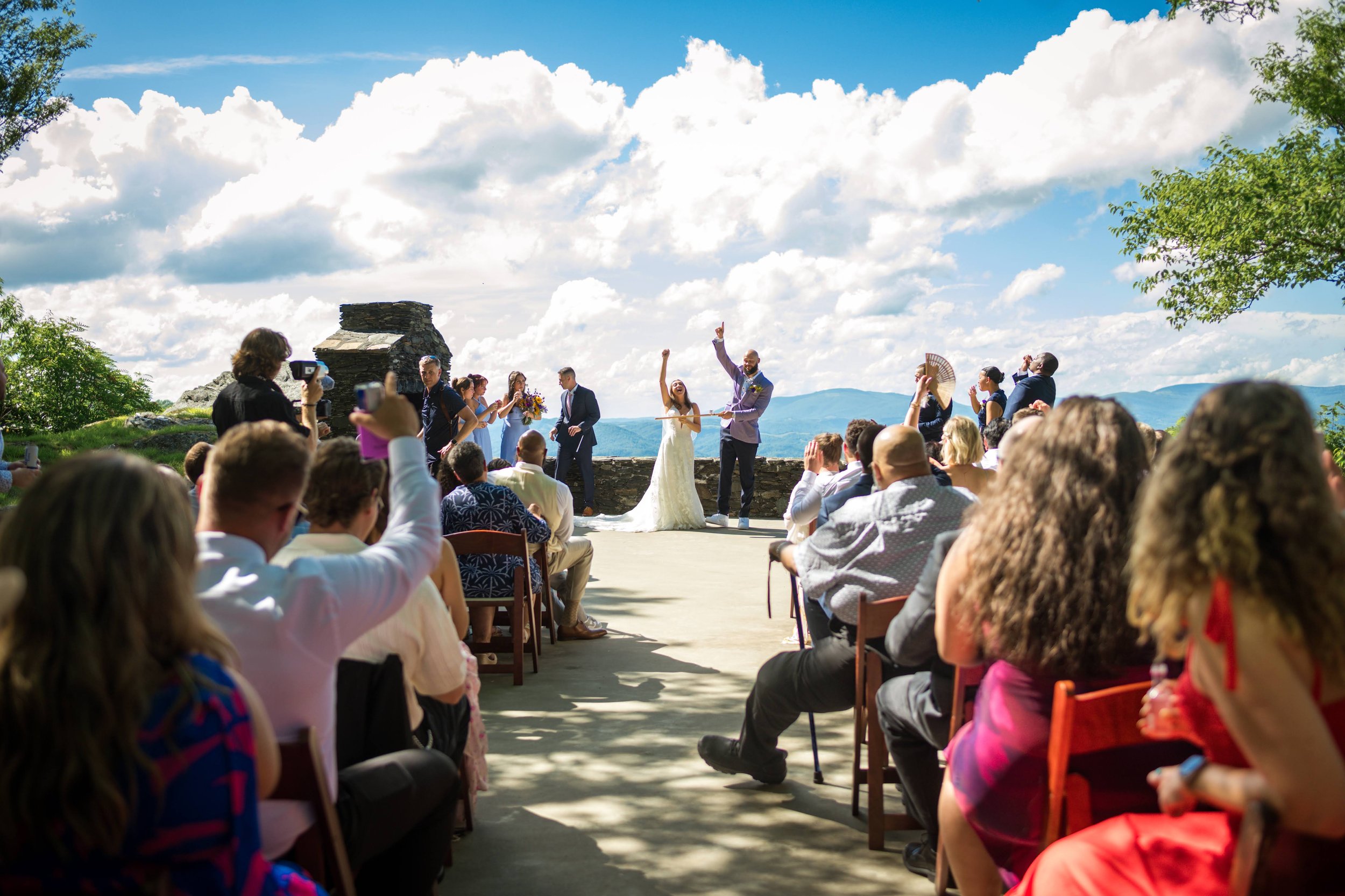 Outdoor wedding ceremony with guests celebrating the bride and groom against a scenic mountain view and bright cloudy sky